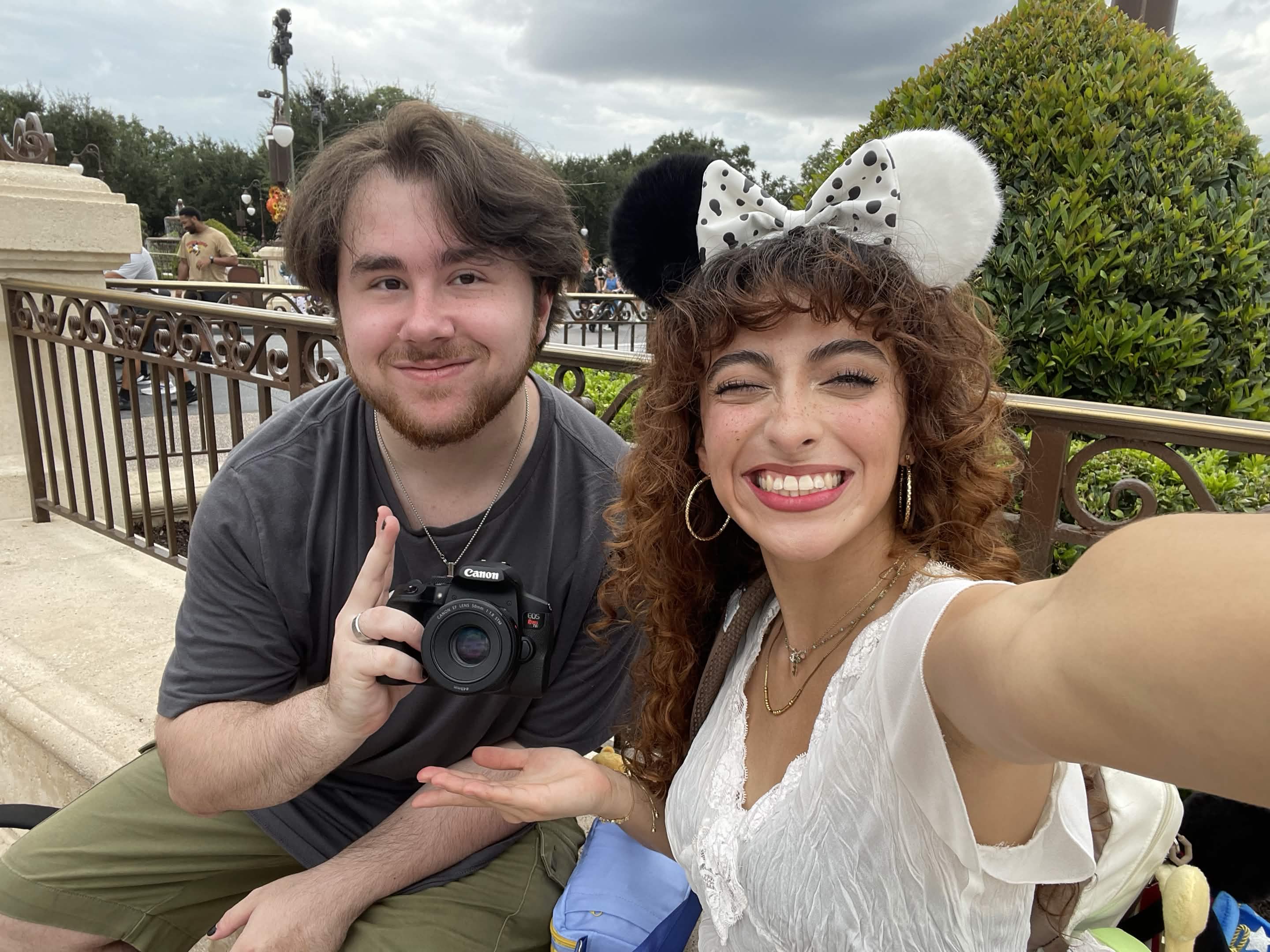 Two people smiling outdoors, one wearing Minnie Mouse ears, the other holding a camera, with a railing and greenery in background.