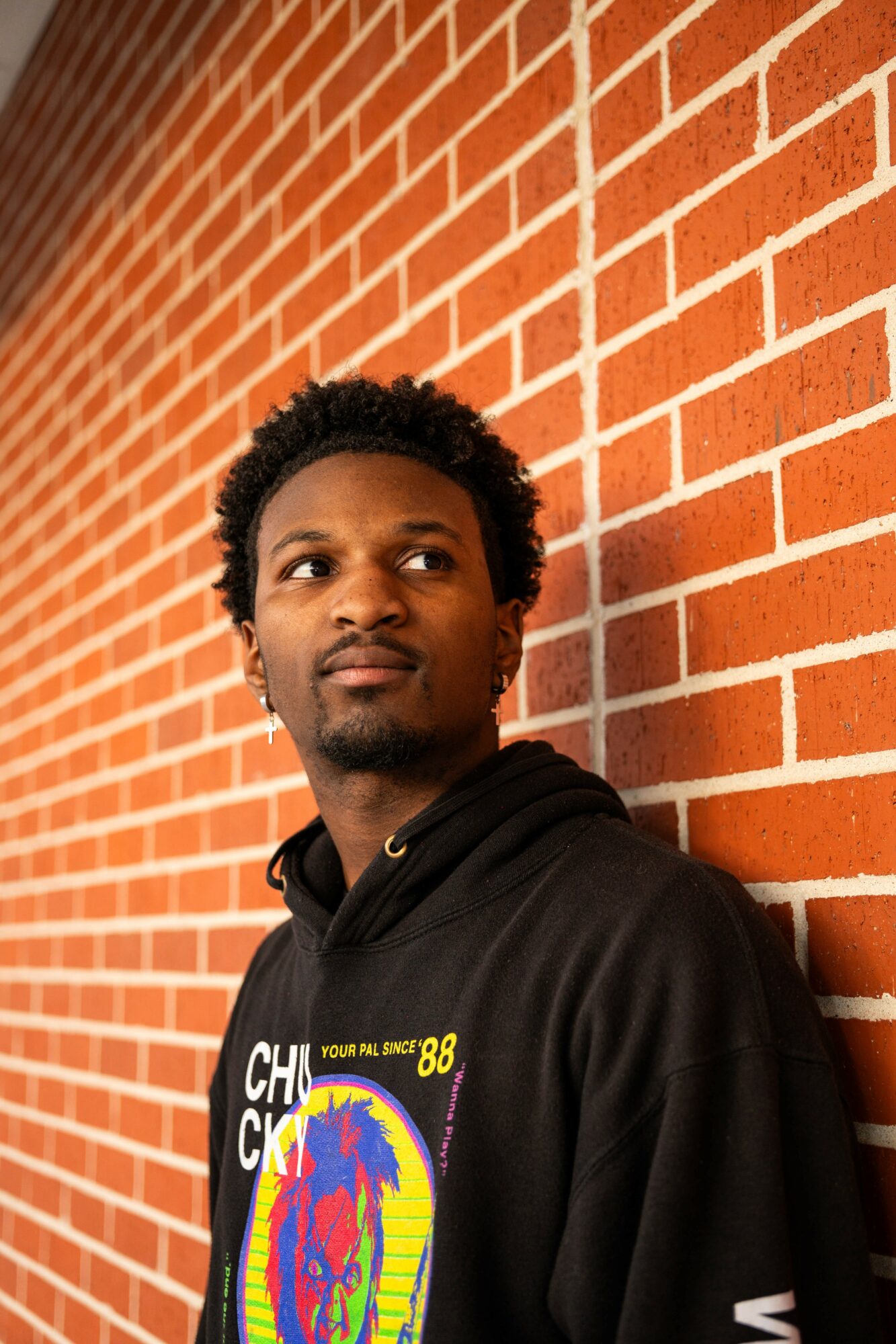 Young man with curly hair leaning against a red brick wall, wearing a black hoodie with colorful graphic.