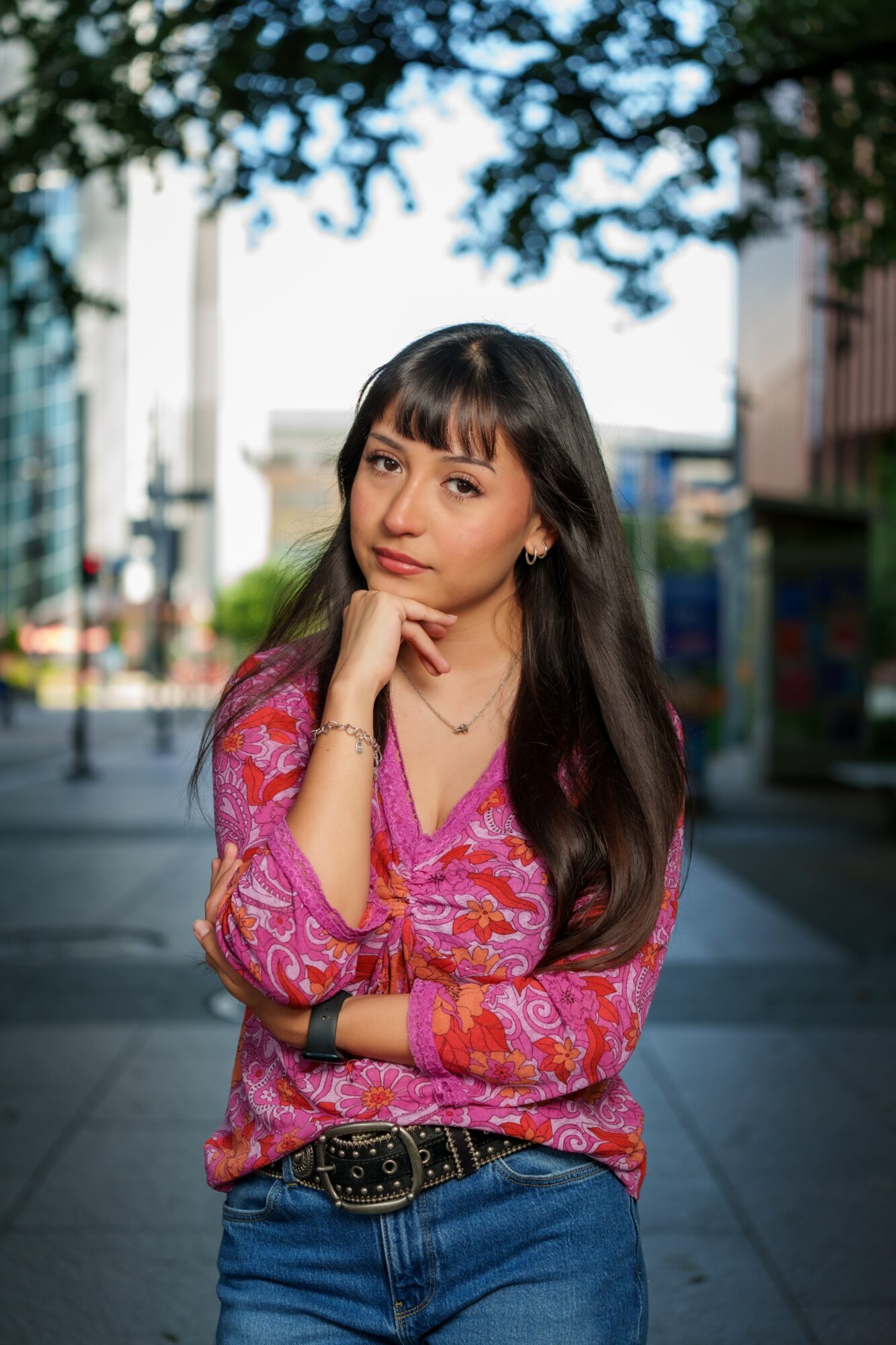 Young woman with long dark hair and bangs, wearing a pink patterned top and blue jeans, standing outdoors with trees and buildings behind her.