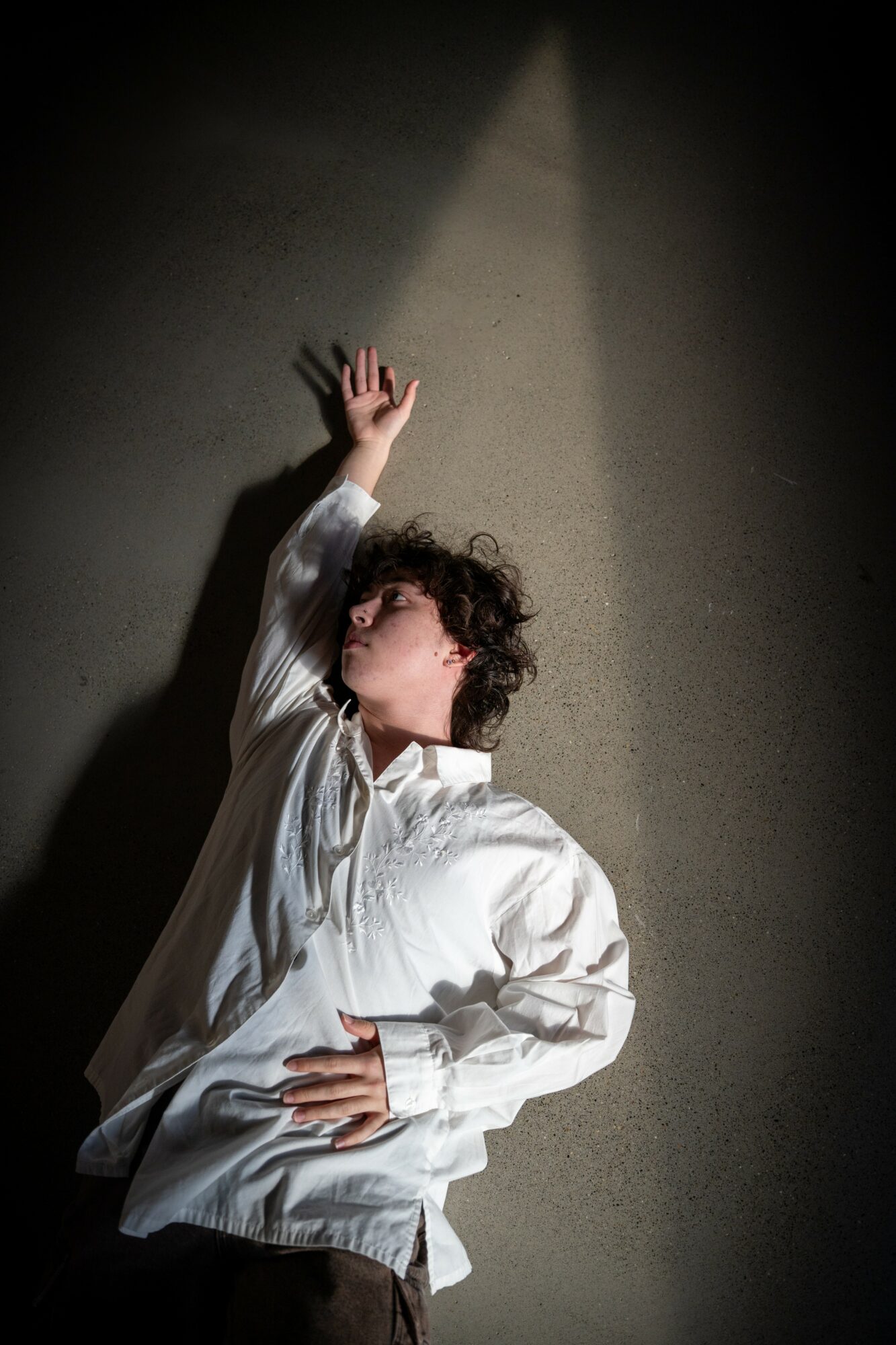 Person with curly hair in a white shirt lying on the floor, reaching upward with one hand, shadow cast on wall.