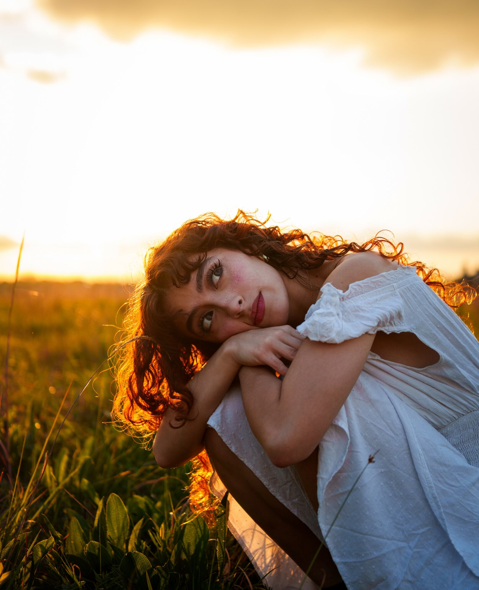 Young woman with curly hair resting her head on her arms in a field during sunset, wearing a white dress.