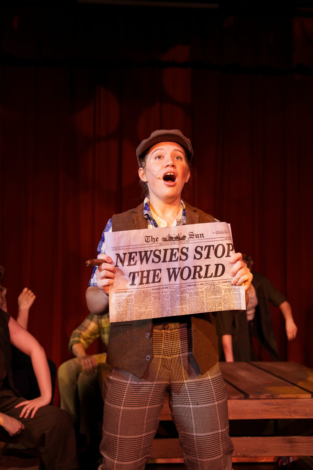 Person on stage holding a sign that reads 'NEWSIES STOP THE WORLD' with a red curtain background.