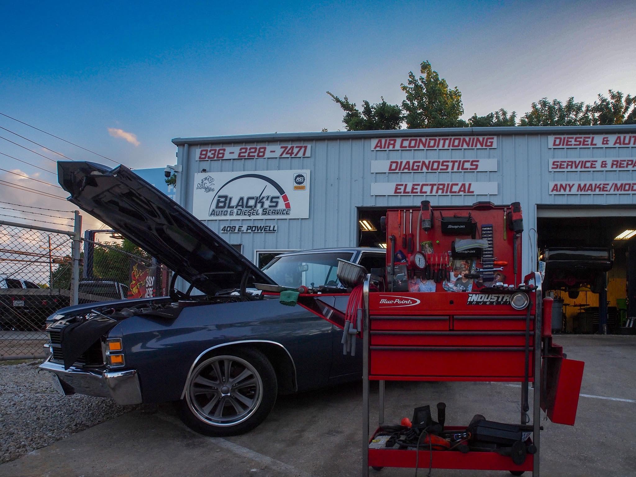 Black classic car with open hood parked in front of a white service truck, red tool chest nearby, outdoors during daytime.