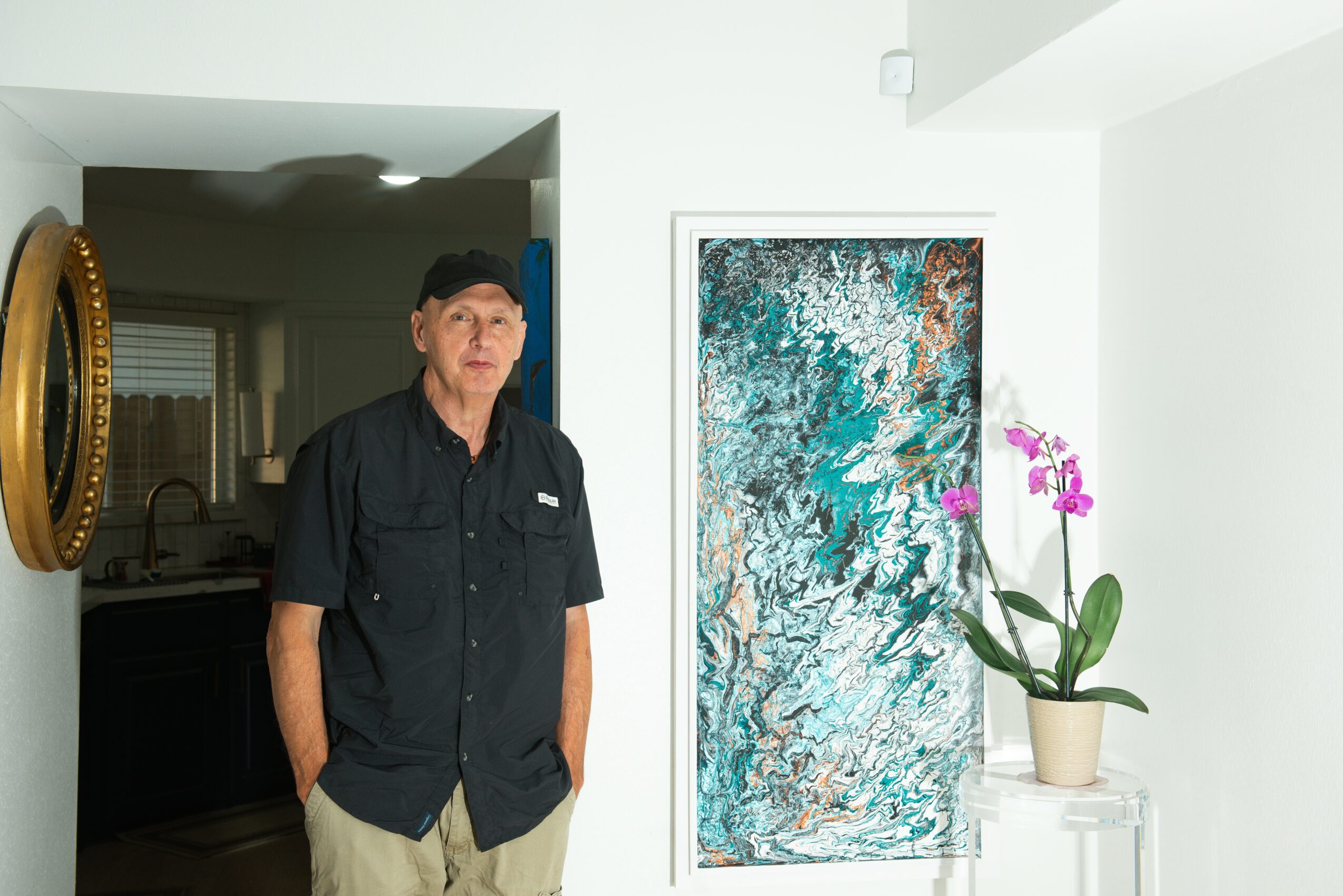 Man in black shirt and cap standing indoors near a colorful abstract painting and a potted orchid, with a kitchen in the background.