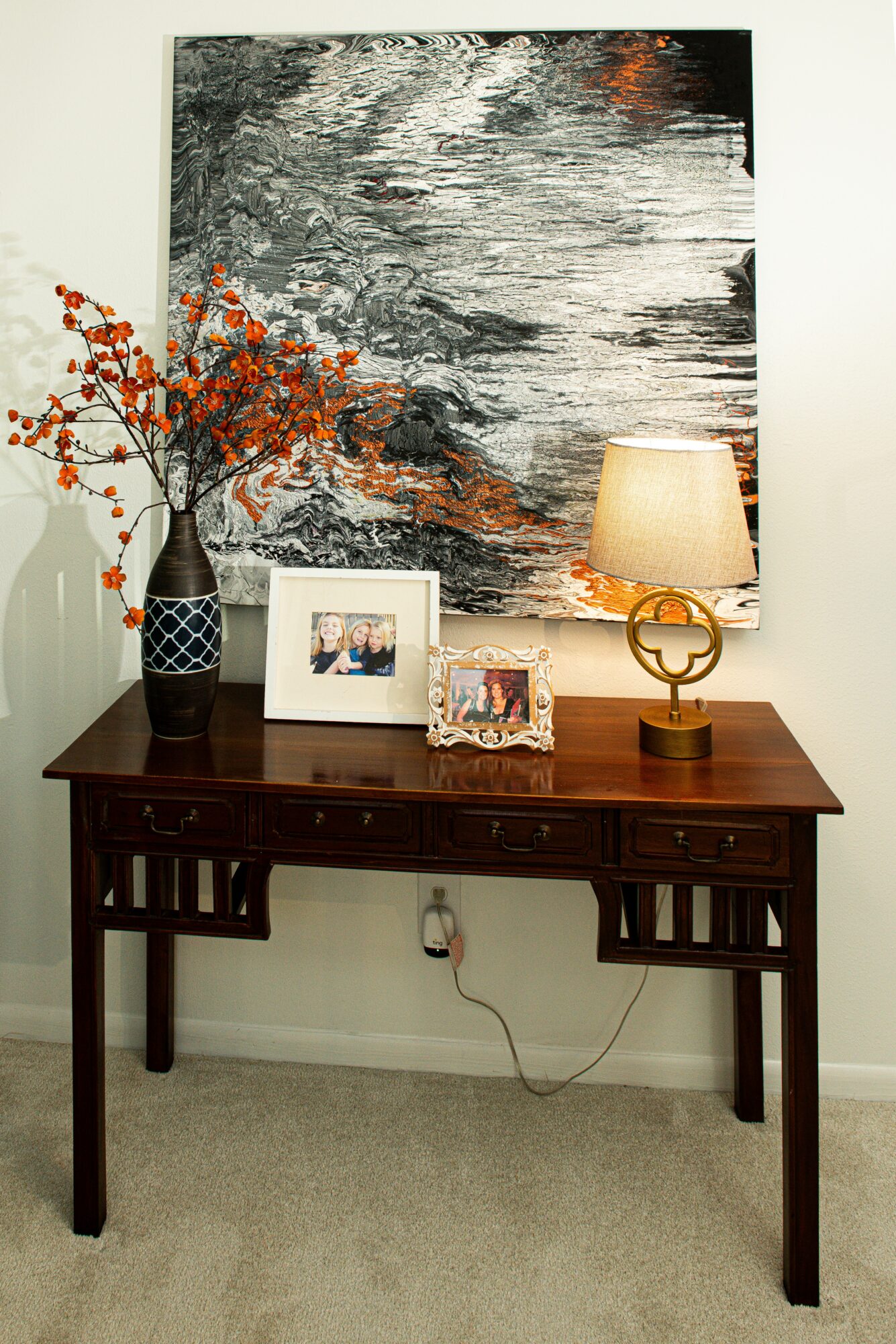 Decorative table with a vase of orange berries, framed photos, and a lamp, against a wall with abstract black and white art.