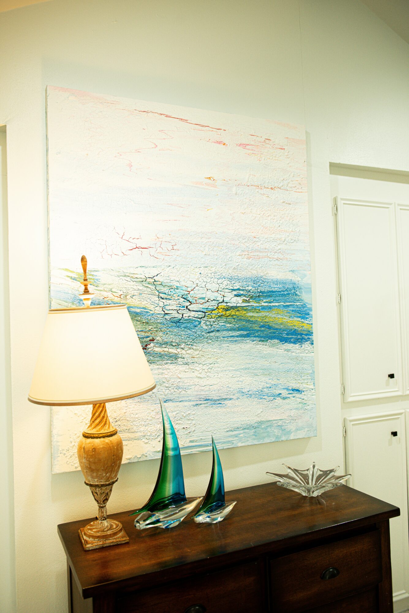 Interior scene with a large abstract painting, a table lamp, and decorative glass objects on a wooden table.