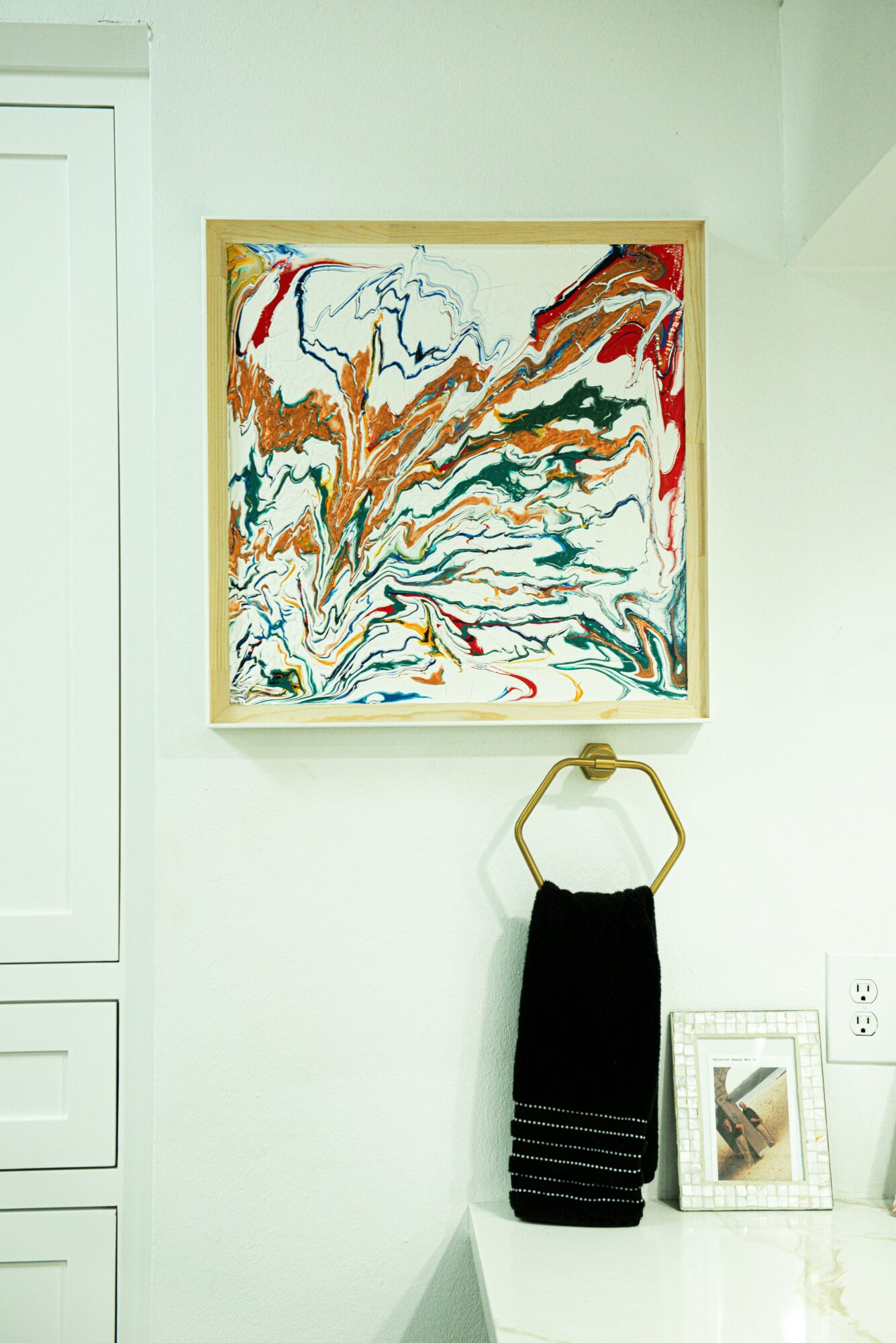 Abstract colorful painting on wall, with black towels hanging on a gold ring, and a framed picture below.