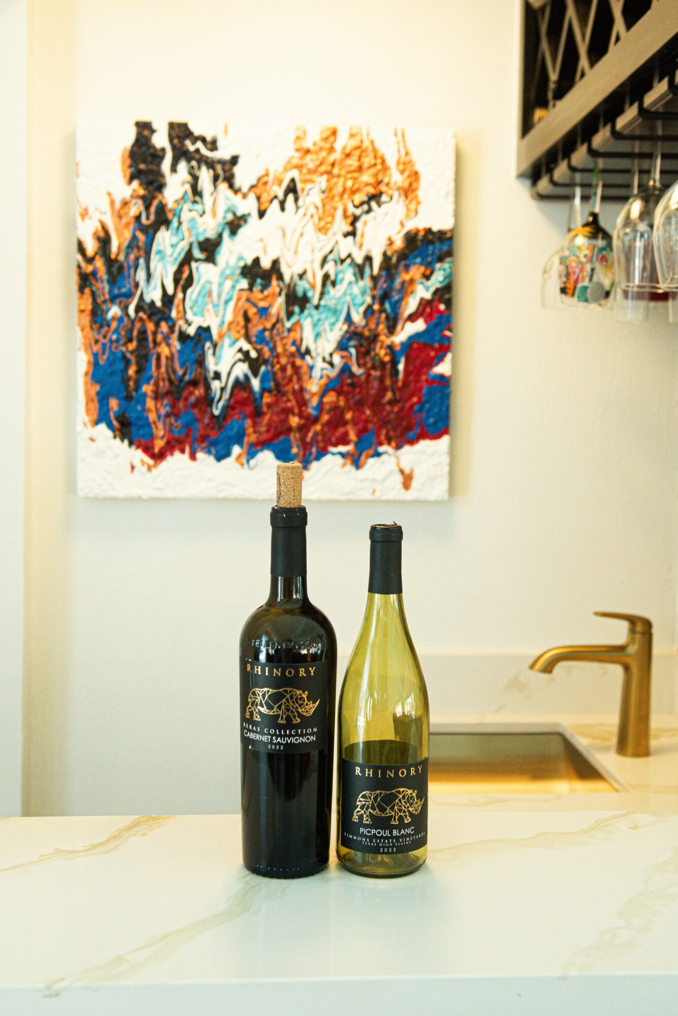 Two bottles of wine on a kitchen counter with a colorful abstract painting on the wall behind, and glasses hanging above.