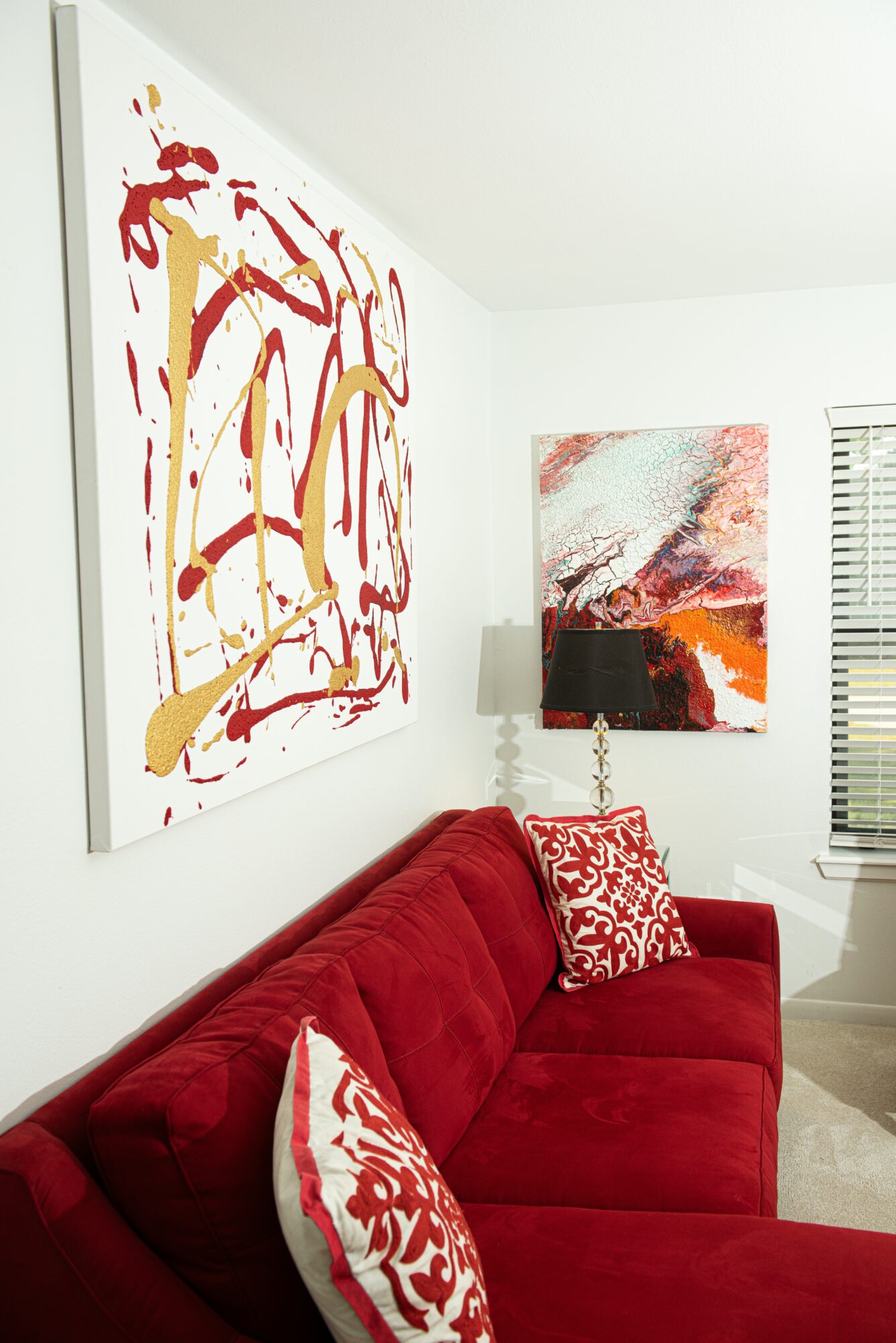Living room with red sofa, abstract paintings on white walls, and a window with blinds.