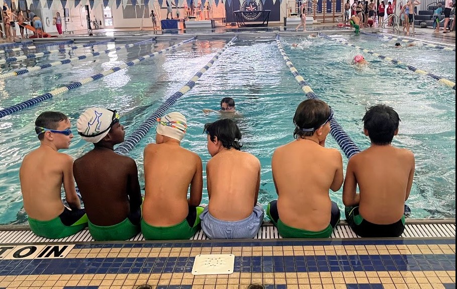 Seven children sit at the edge of a swimming pool, watching a swim practice or competition, with swimmers in the water.