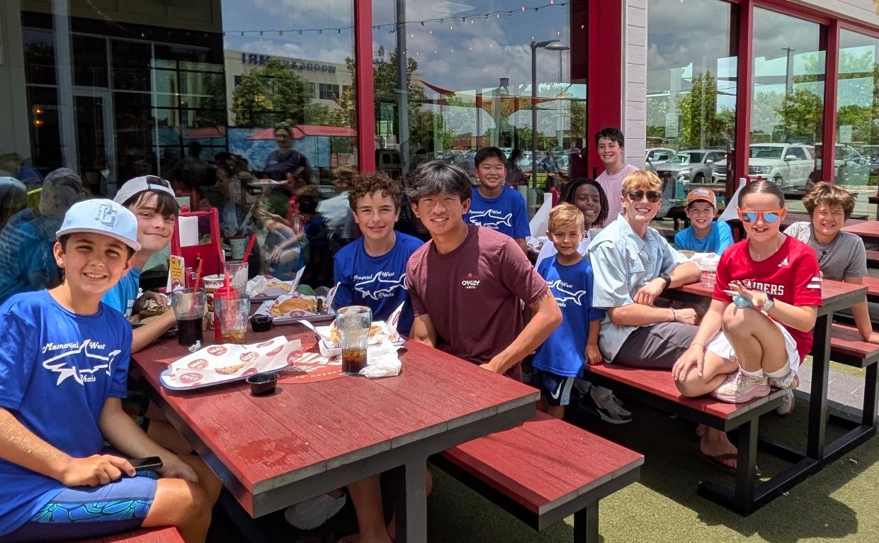 Group of children and teenagers sitting at outdoor tables, smiling, with a building and glass windows in the background.
