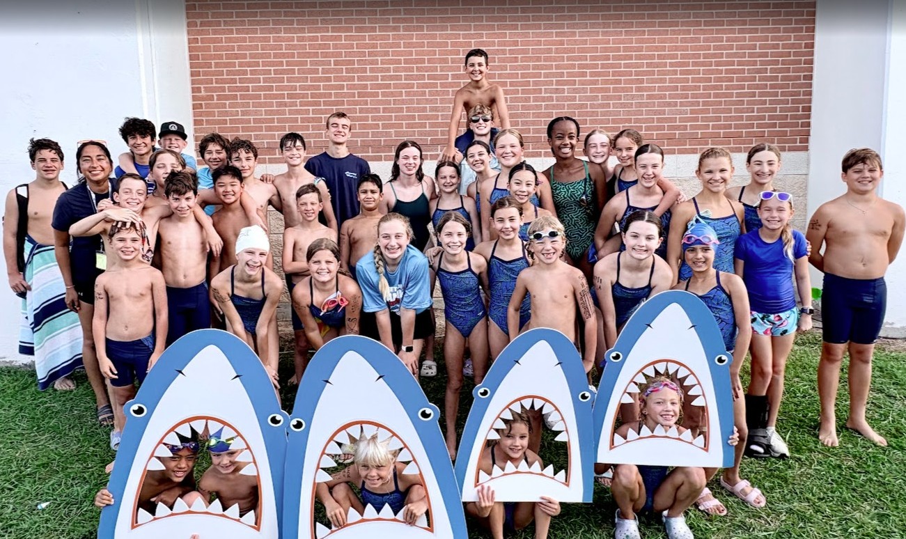 Group of children and teenagers in swimsuits outdoors, some behind shark-shaped pool toys, smiling and posing for a photo.