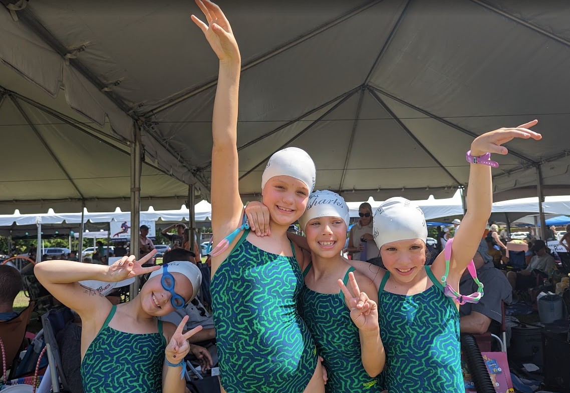 Four young girls in matching swimsuits and swim caps smiling and making peace signs under a tent.