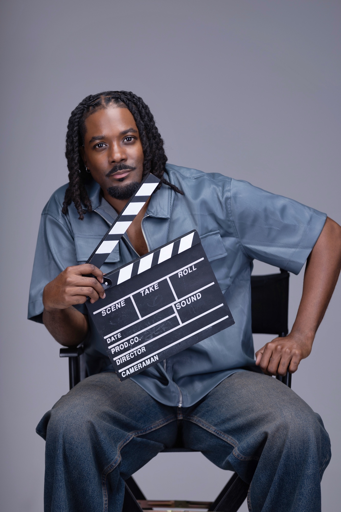 Person with dreadlocks holding a film slate, sitting on a chair against a plain background.