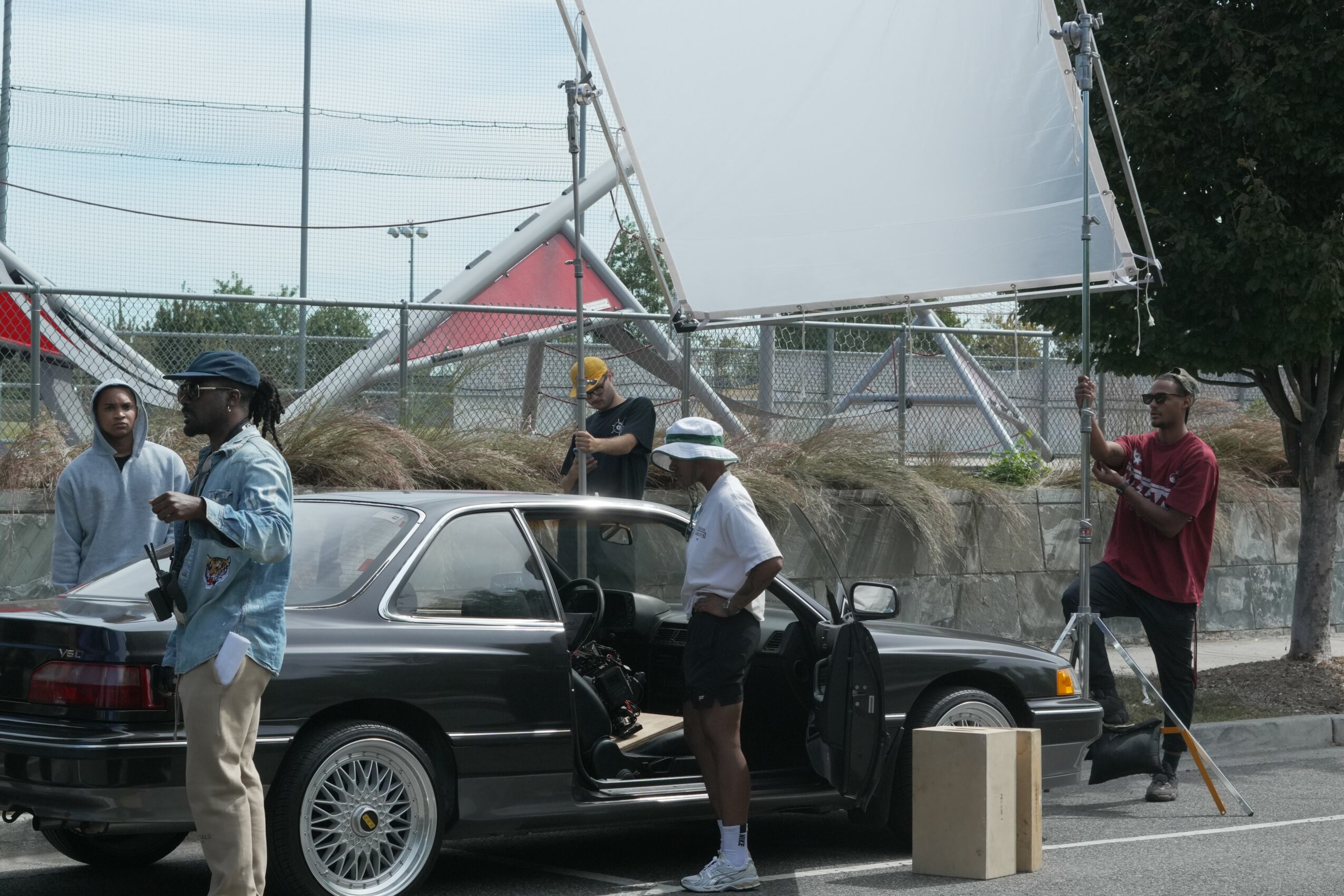 Group of people around a black car with open door, some wearing hats, in an outdoor area with trees and structures.