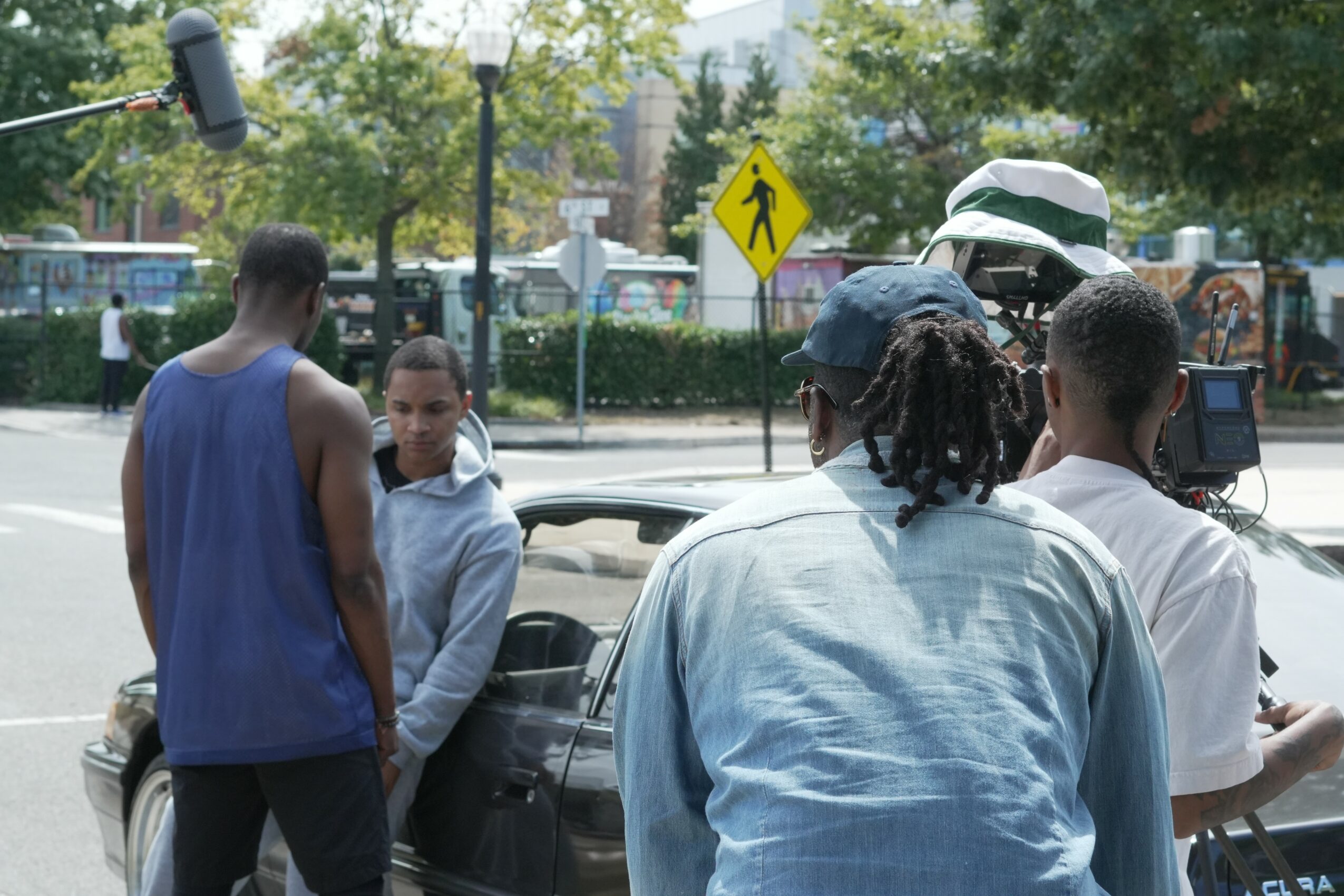 Group of four people standing near a car on a city street, with trees and a pedestrian crossing sign in background.