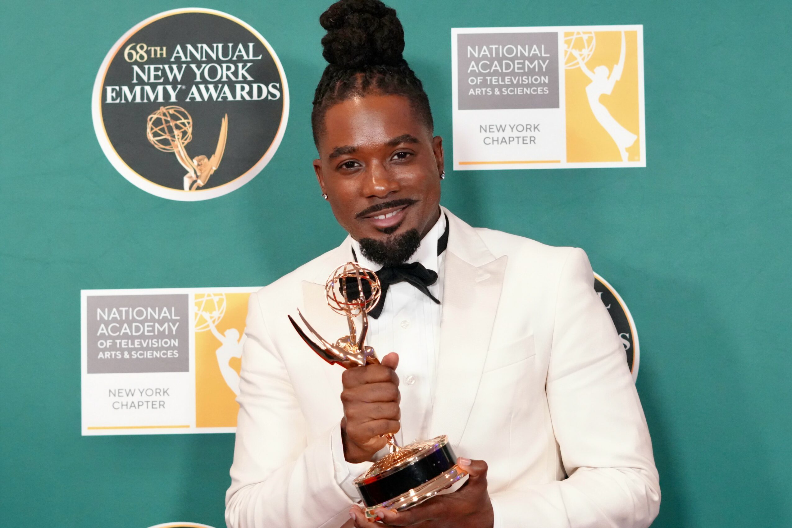 Person in white tuxedo holding an Emmy award, standing in front of a teal background with event logos.