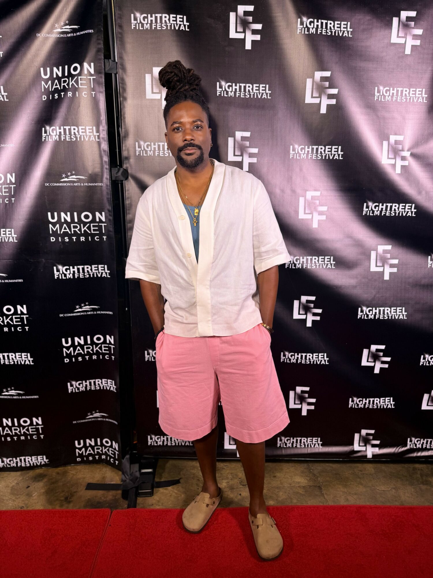 Man with a beard and bun hairstyle standing on red carpet in front of black backdrop with logos, wearing a white shirt and pink shorts.