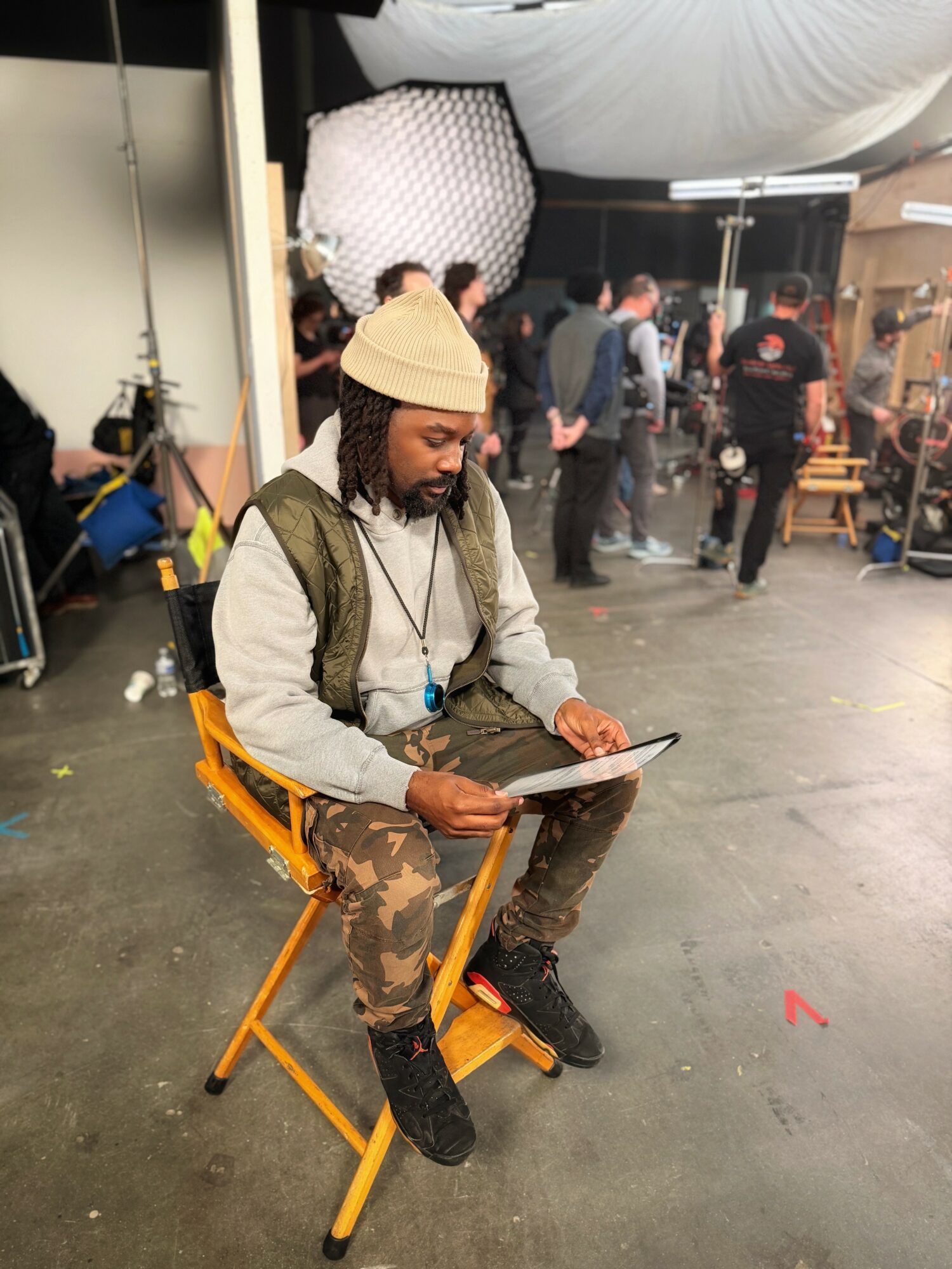 Man with a beige beanie and backpack sitting on a yellow folding chair, writing on a notepad in a busy indoor setting.