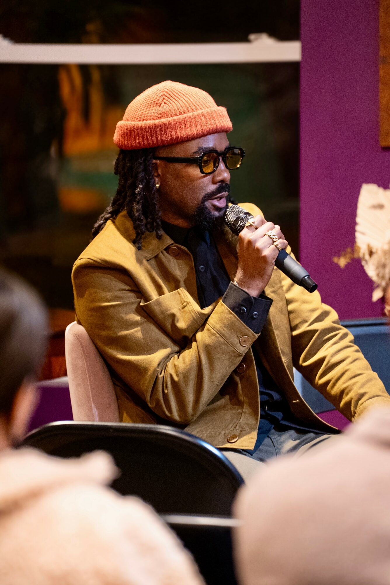 Man with glasses and a beanie speaking into a microphone, seated indoors with purple walls.