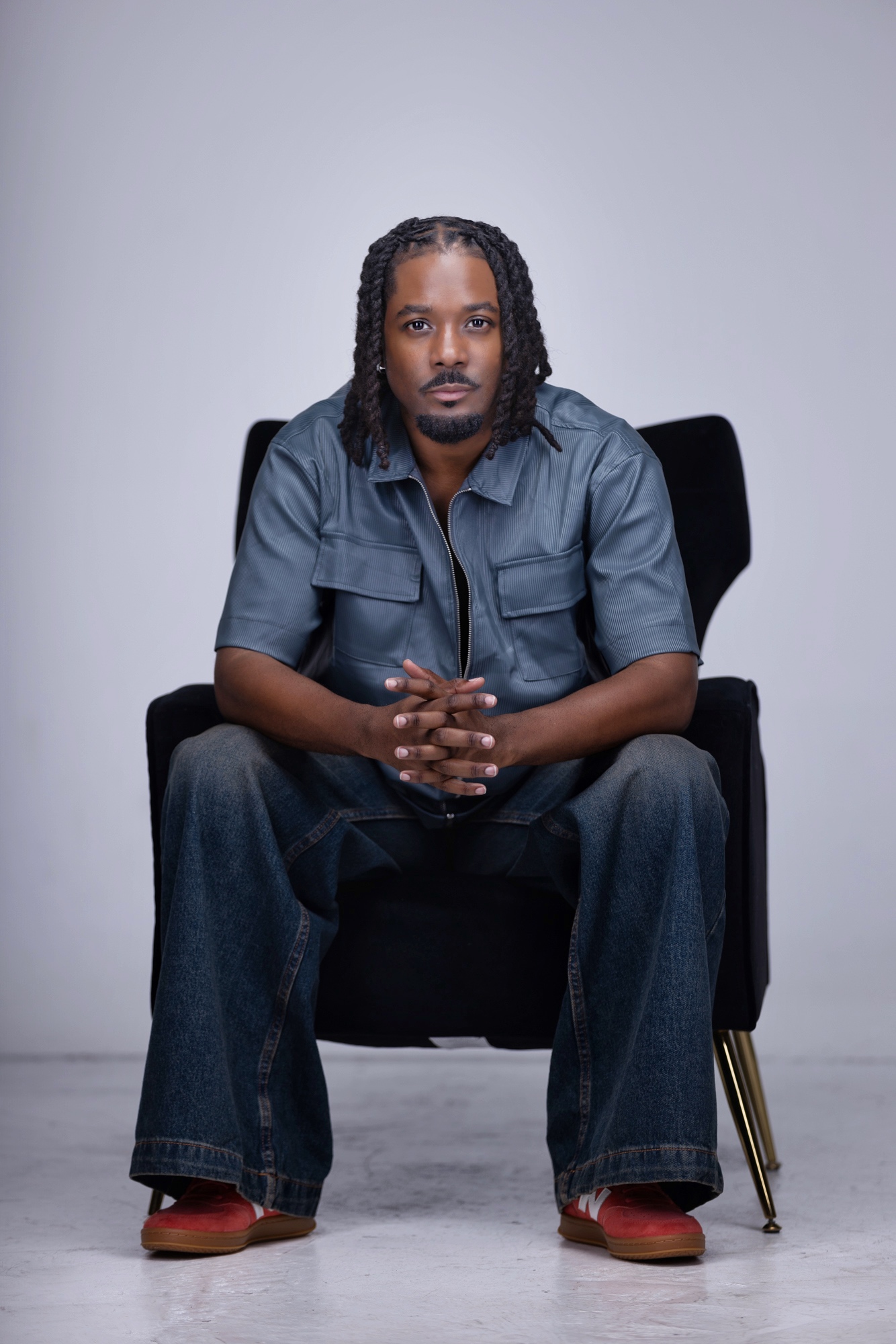 Man with dreadlocks sitting on a black chair, hands clasped, wearing a blue shirt and jeans, against a plain background.