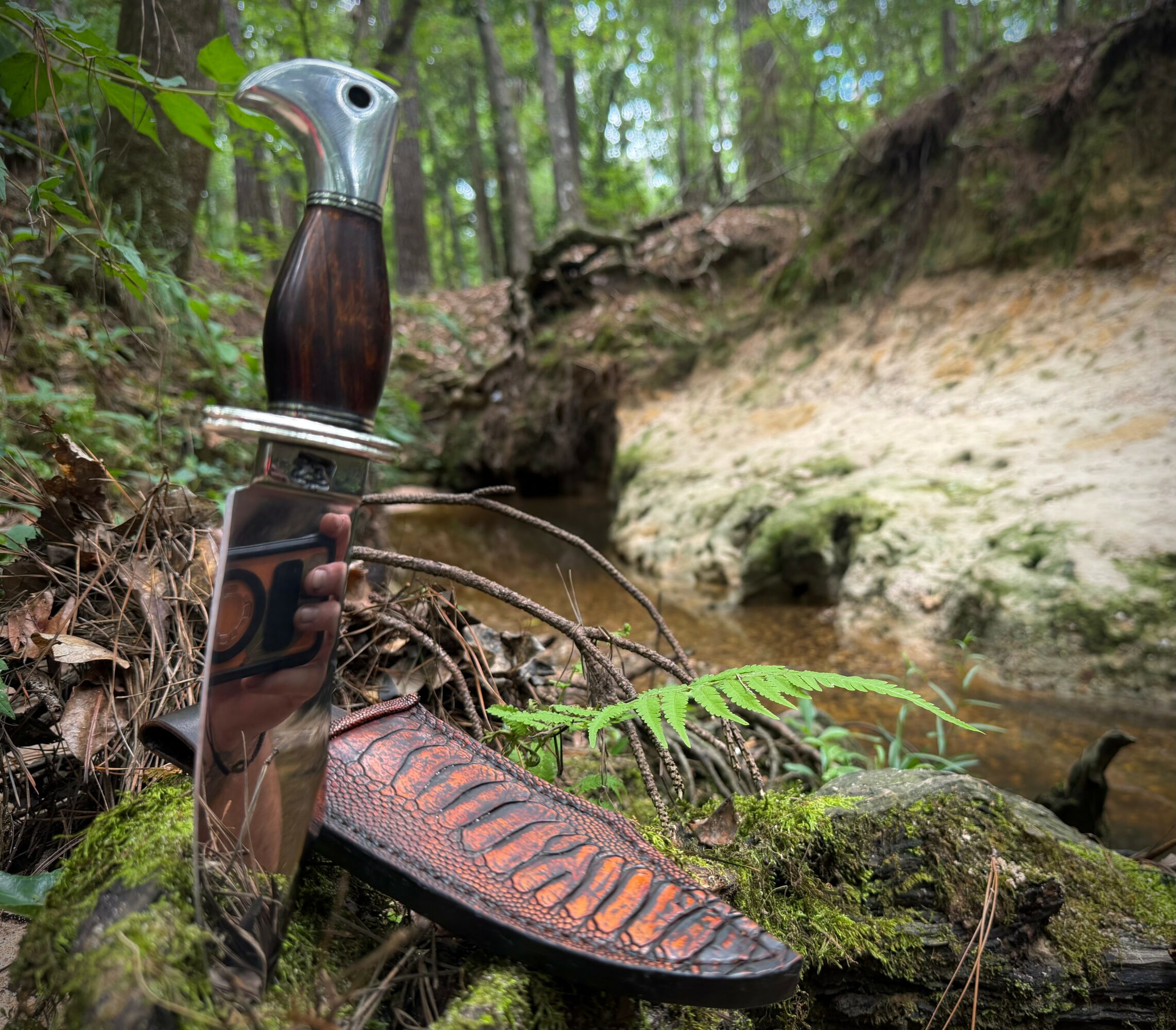 A knife with a wooden handle and decorative blade is placed on mossy ground near a small stream in a forest.