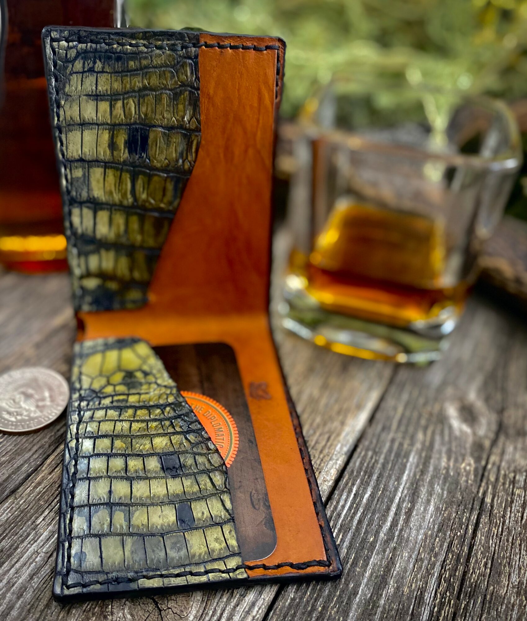 Open wallet with crocodile pattern, placed on a wooden surface with a glass of whiskey in the background.