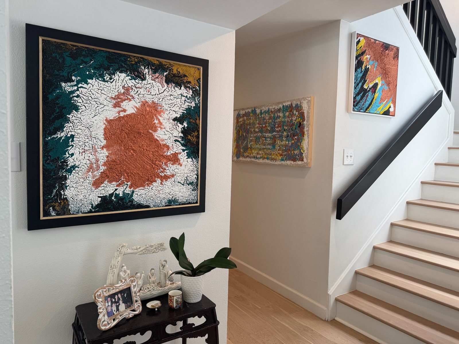 Living room corner with large abstract painting, smaller artwork, staircase, and a table with decorative items and a plant.