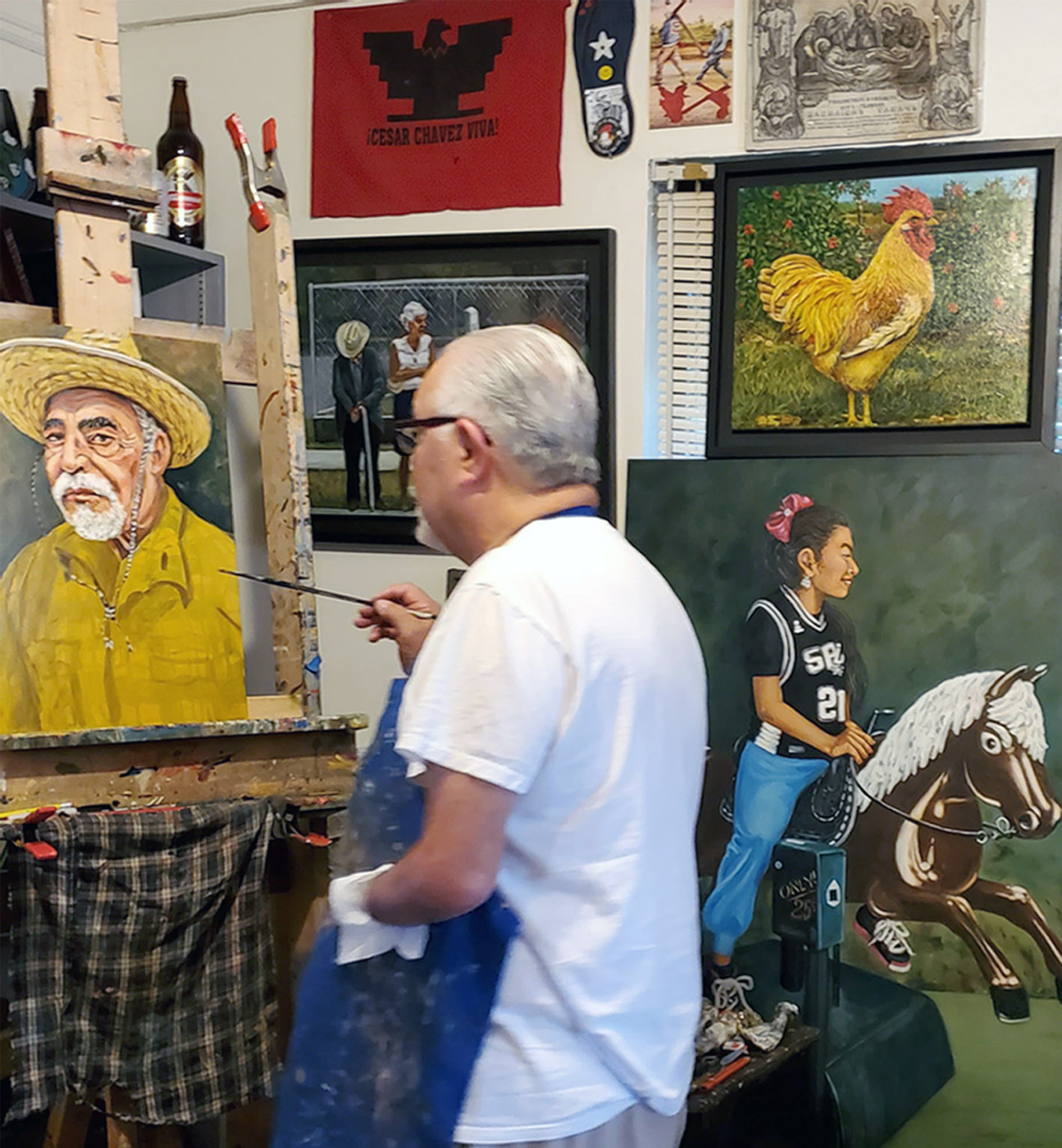 Man with glasses and white shirt painting a portrait of an older man with a hat, surrounded by framed artwork.