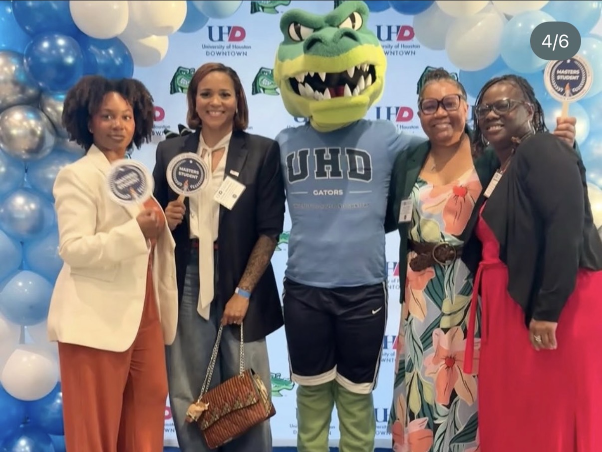 Group of five people, including a mascot with a green head and blue shirt, standing in front of balloons and a backdrop.