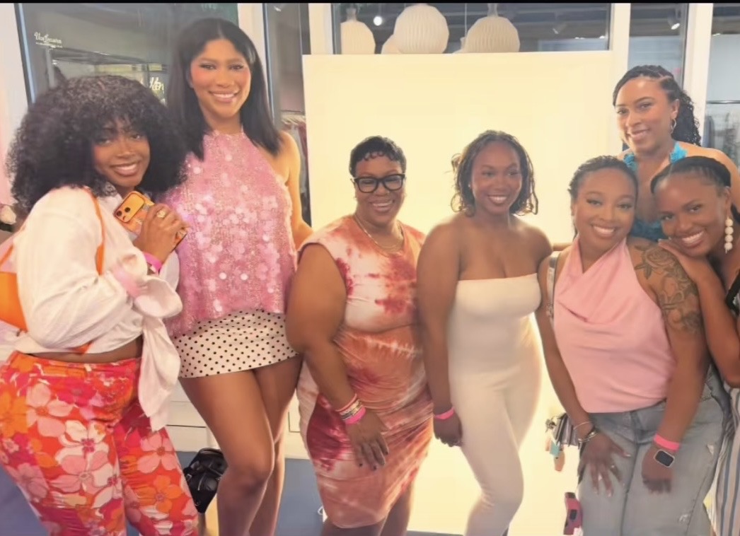 Group of seven women smiling, standing indoors in front of a white background, wearing colorful outfits.