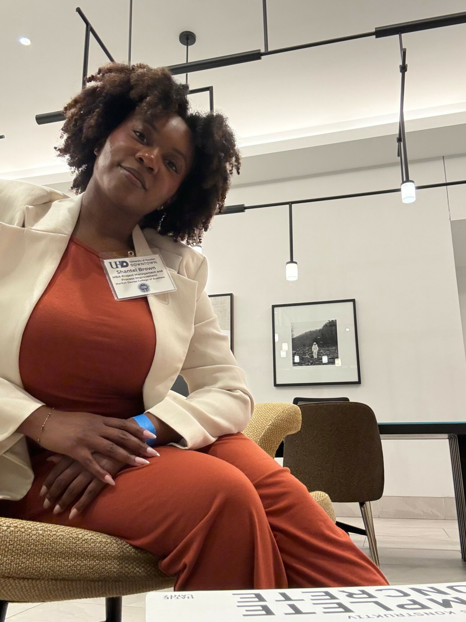 Woman with curly hair sitting in a chair, wearing a beige blazer and rust-colored dress, in a modern room with framed pictures.