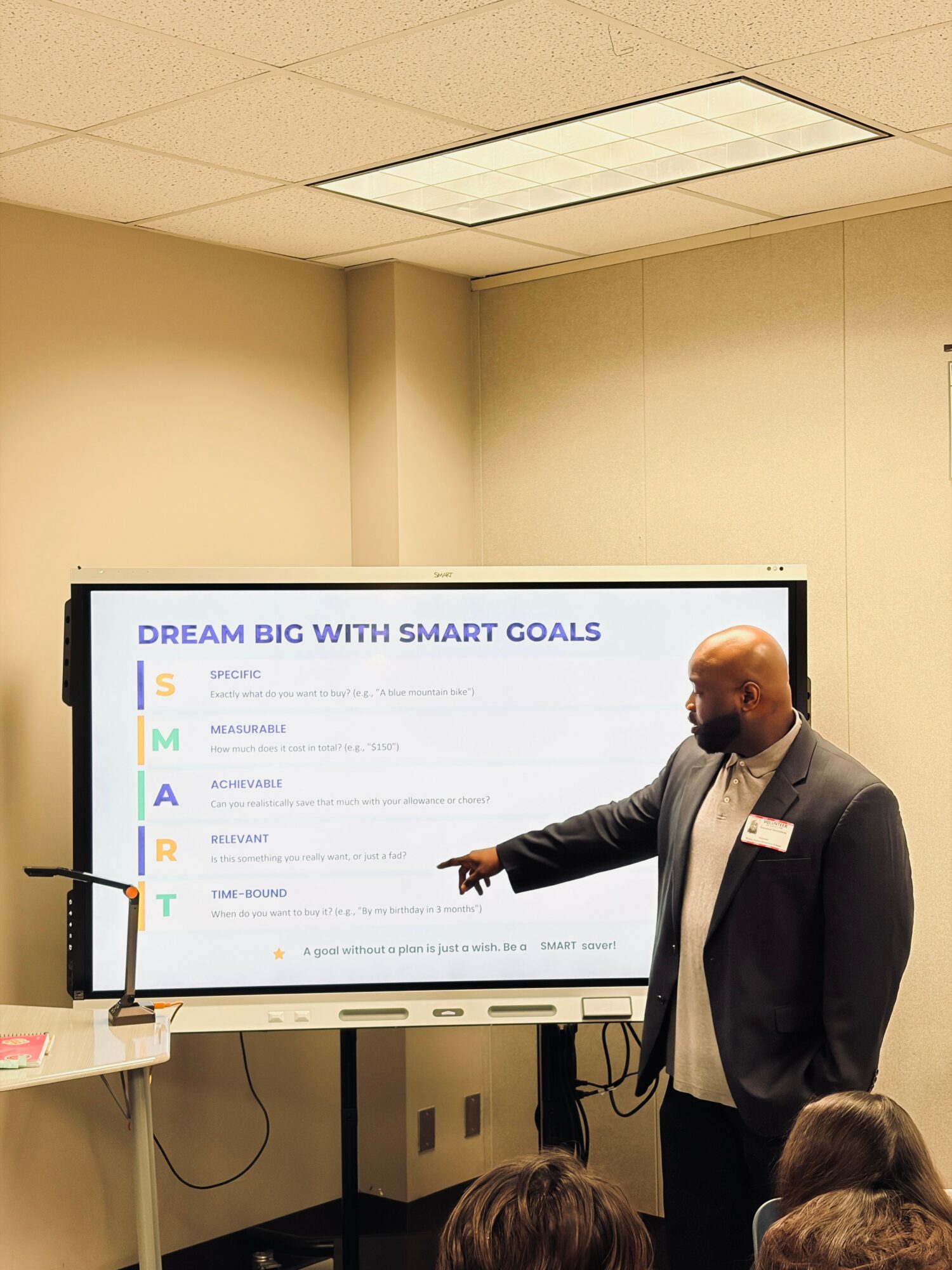 Man in suit and mask pointing at a large screen with SMART goals, audience visible in foreground.