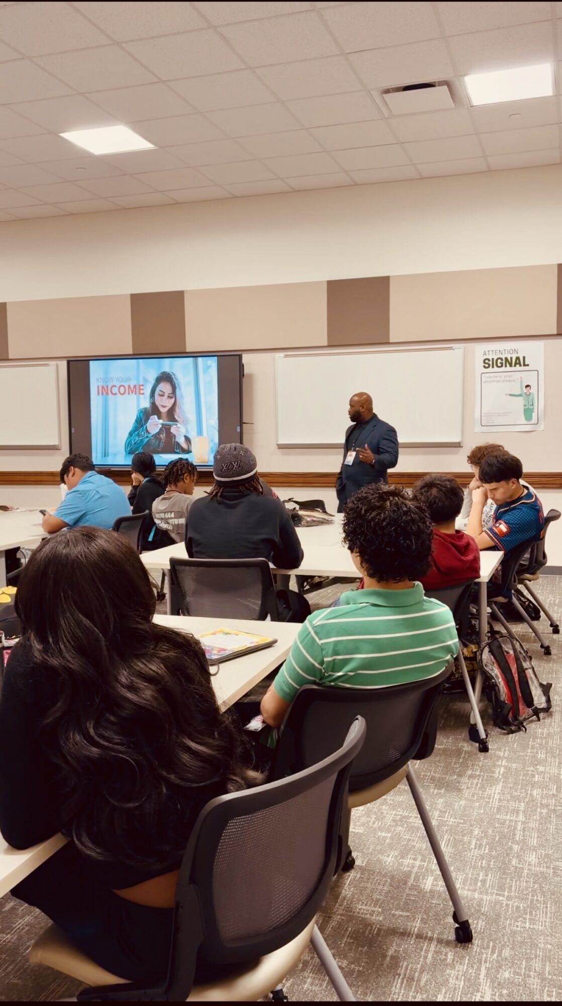 Classroom with students seated at desks, teacher standing near screen, students watching presentation.
