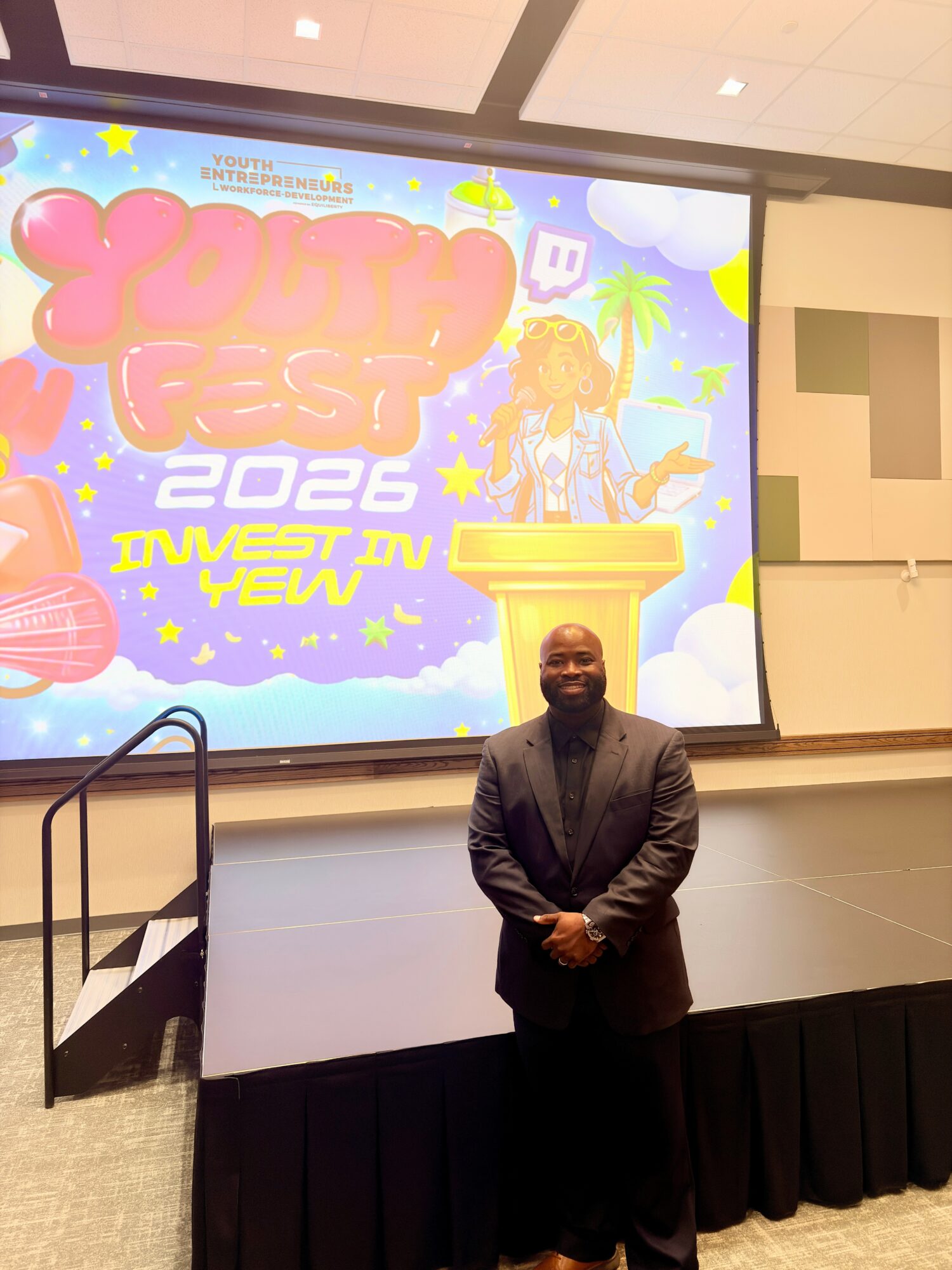 Man in a suit standing on stage in front of a colorful festival poster, smiling, with a podium nearby.