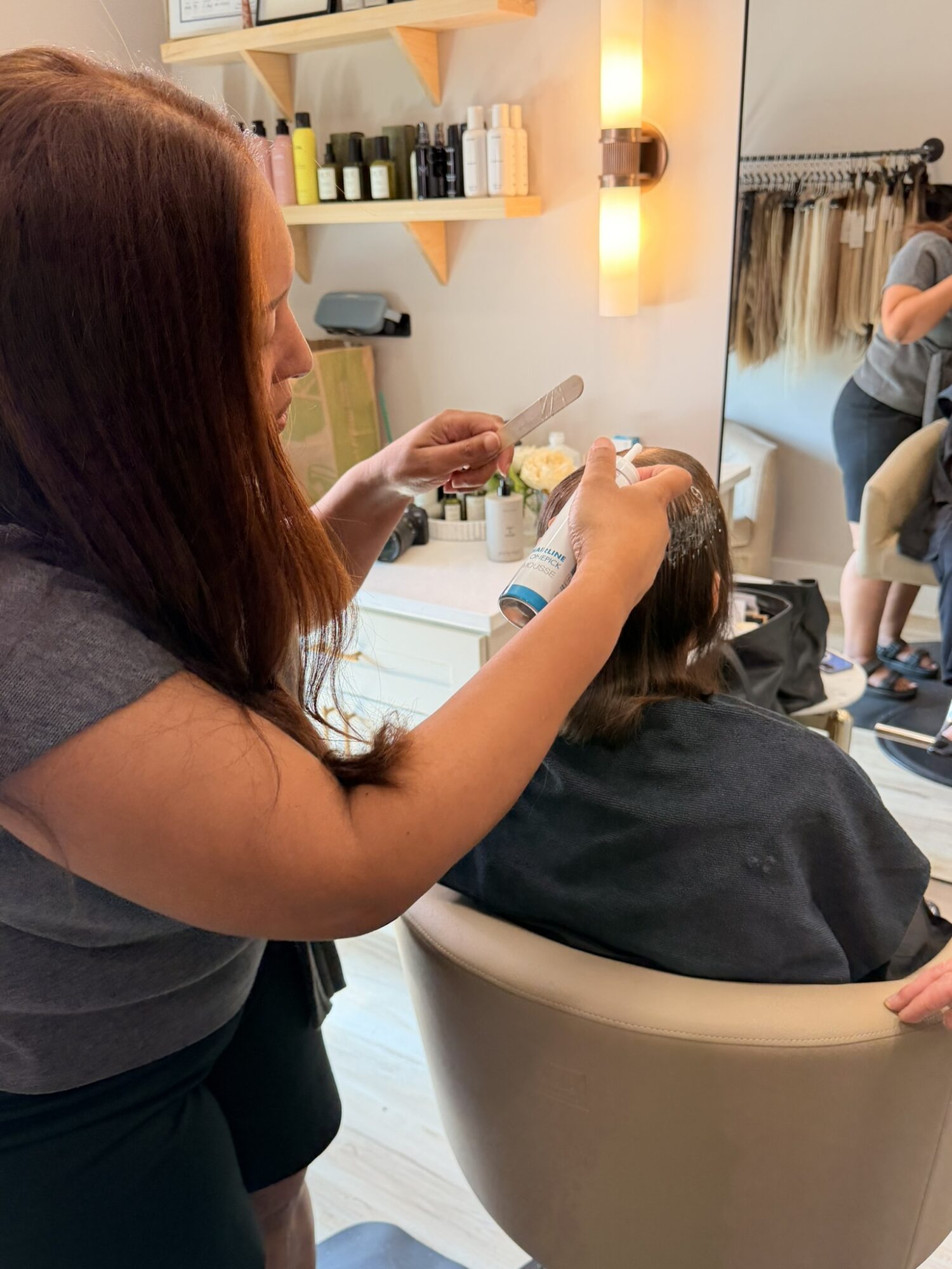 Hairdresser styling a client's hair in a salon with shelves and a mirror, person seated in a white chair.