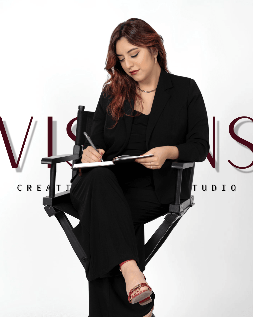 Woman in black suit sitting on director's chair, writing in a notebook, with a white background and partially visible text.