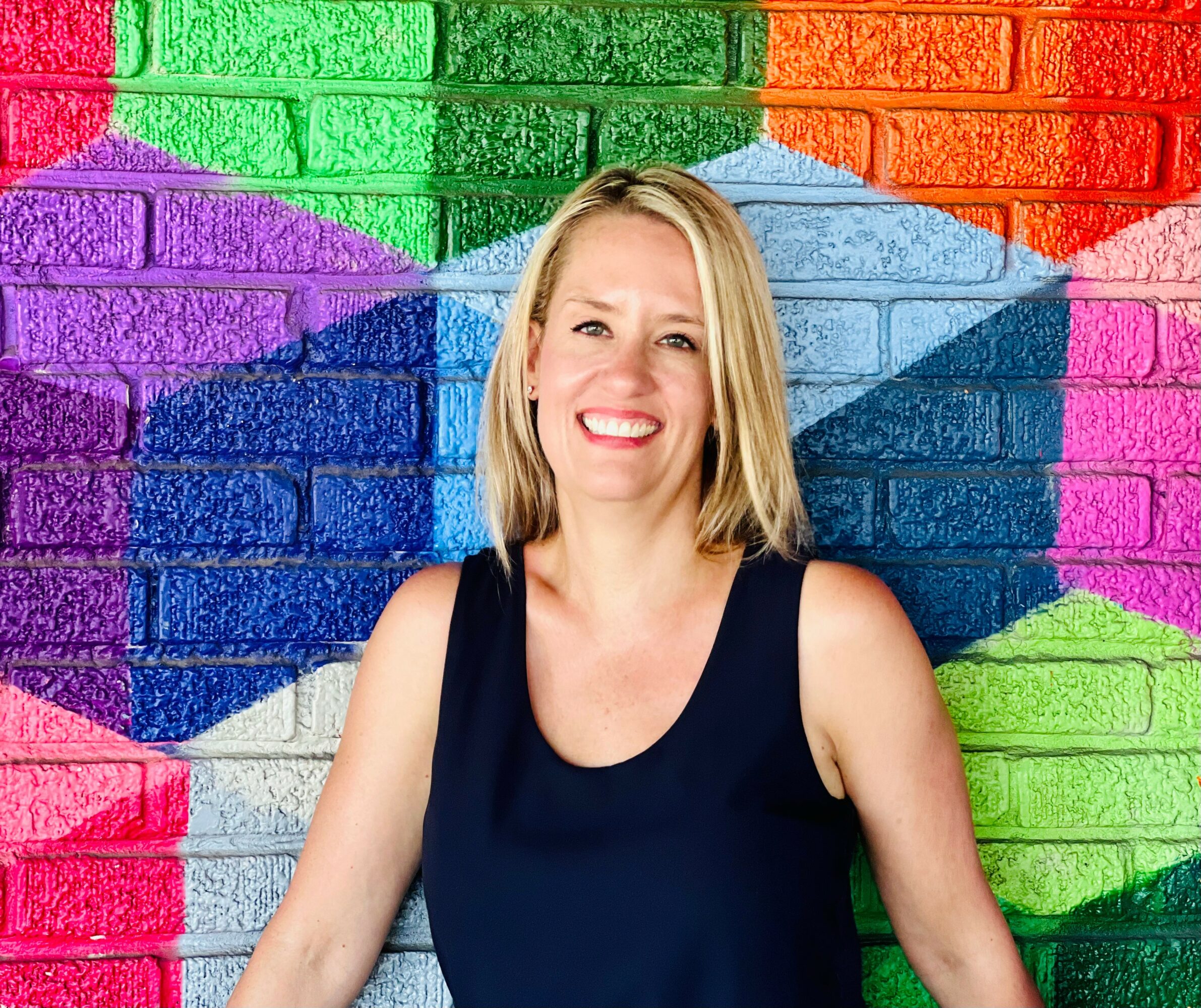 Woman with blonde hair smiling in front of a colorful graffiti wall, wearing a dark sleeveless top.