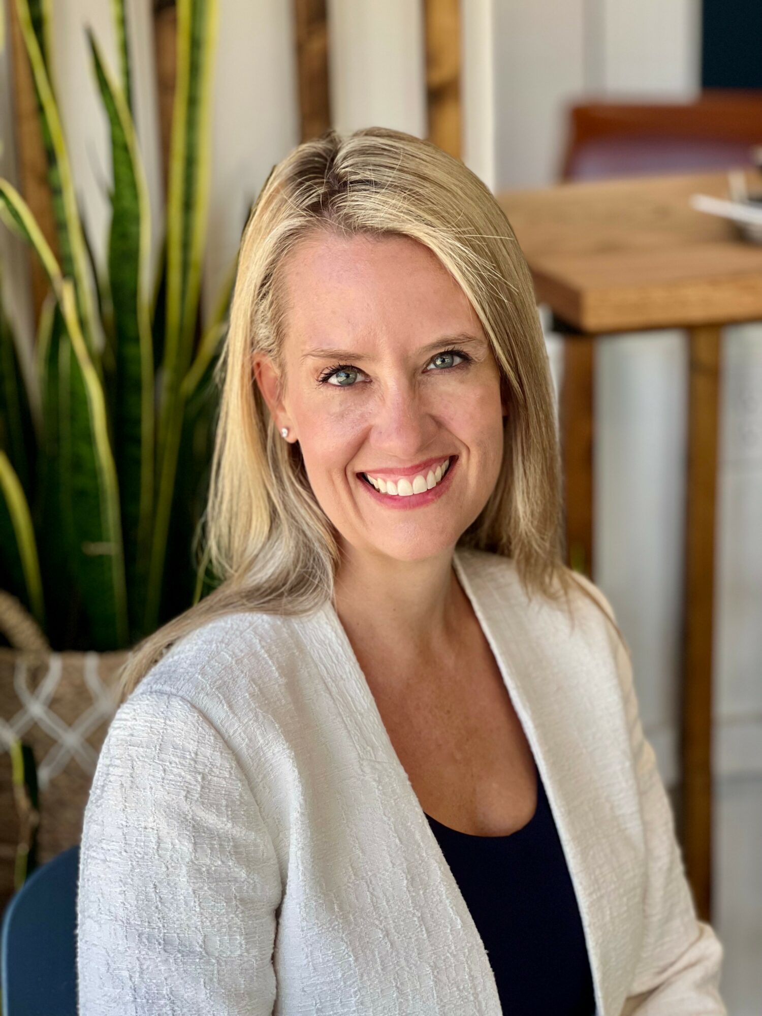 Smiling woman with blonde hair in a white cardigan indoors, with a plant and wooden furniture in the background.
