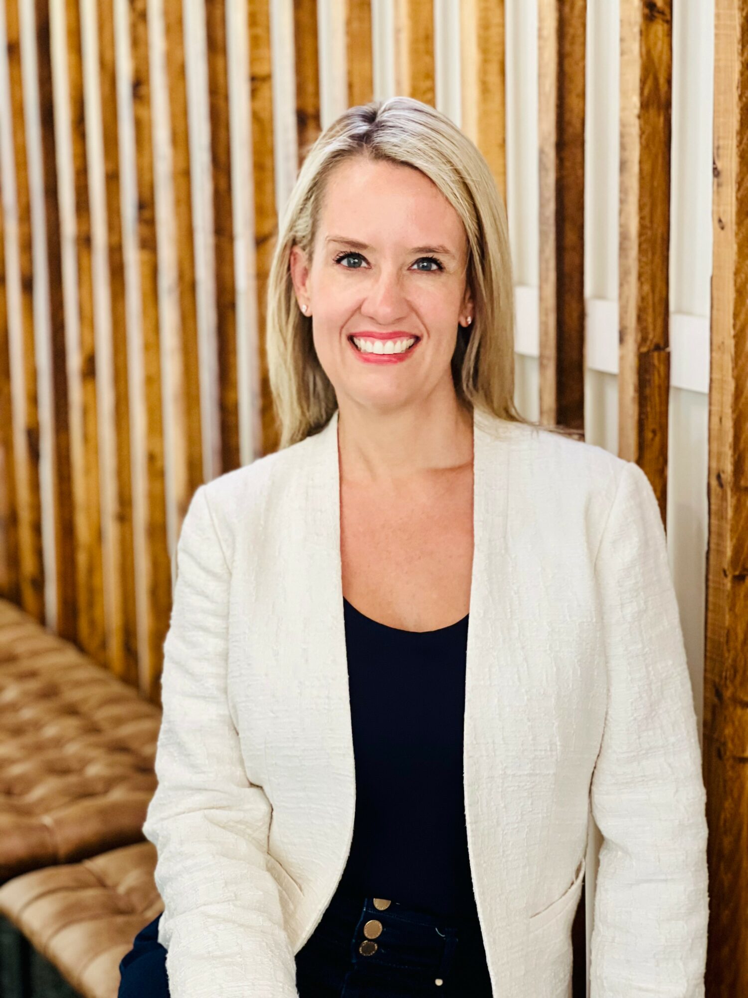 Woman with blonde hair smiling, wearing a white blazer and black top, standing in front of wooden slats.
