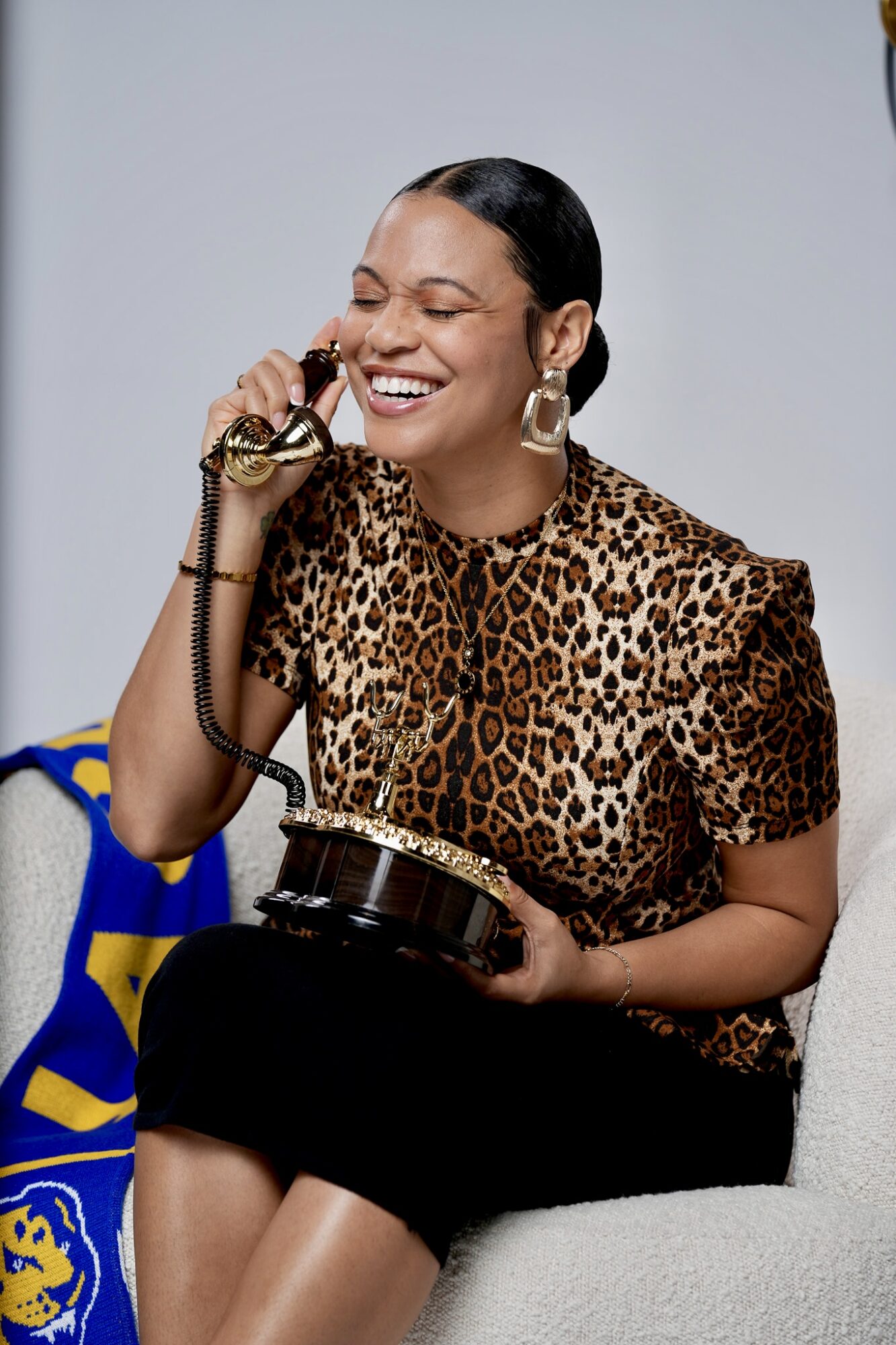 Woman with dark hair in a ponytail, wearing a leopard print top and large earrings, smiling while talking on a vintage telephone.