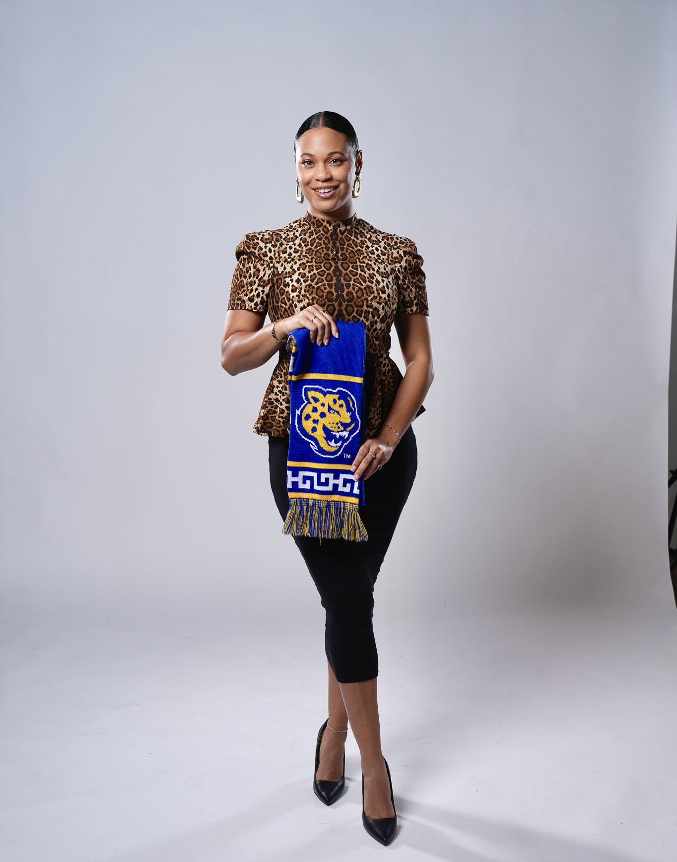 Woman standing and smiling, holding a colorful bag, wearing a leopard print top and black pants, in a studio setting.