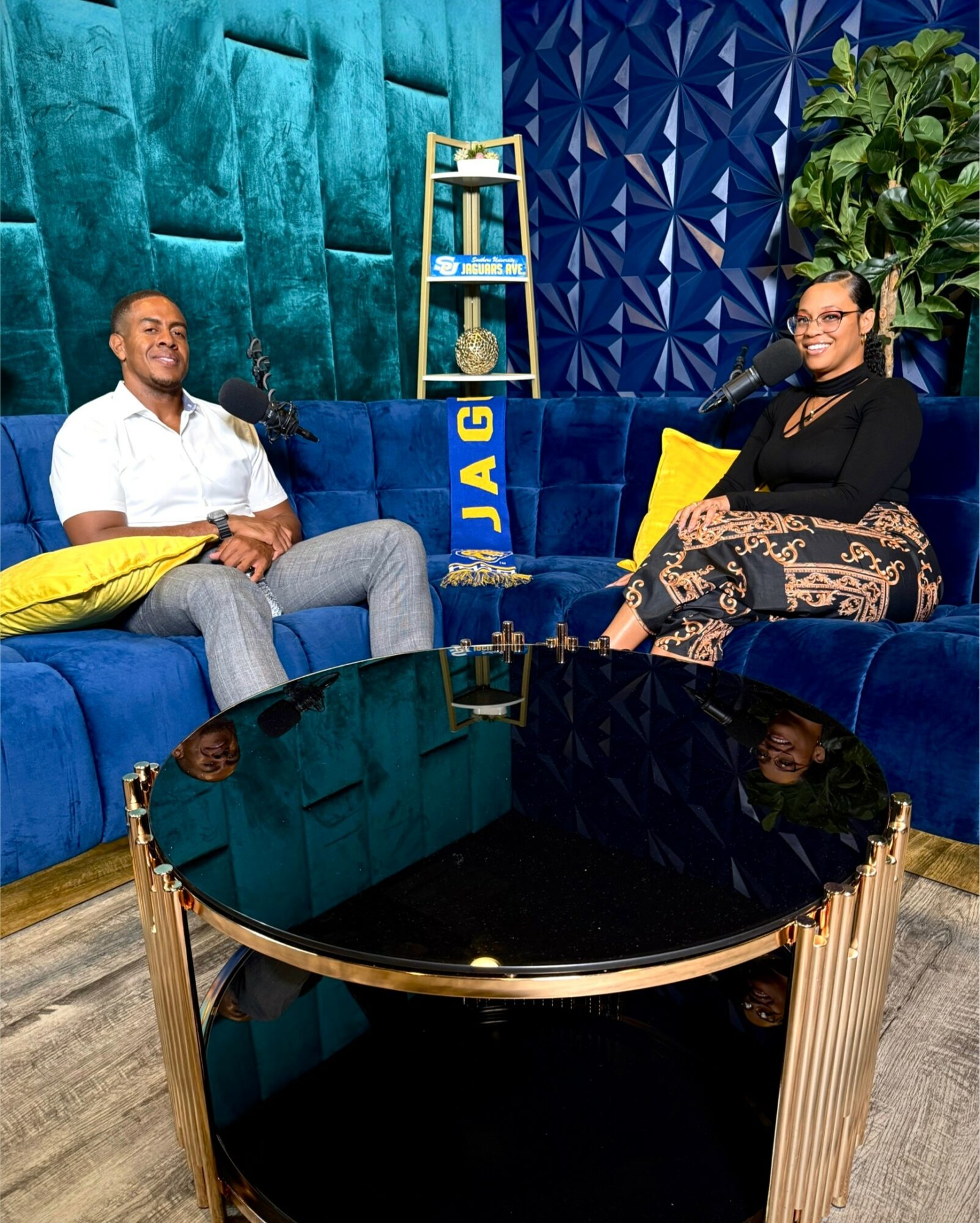 Two people sit on a blue sofa in a colorful room with patterned walls and a large plant, smiling at the camera.