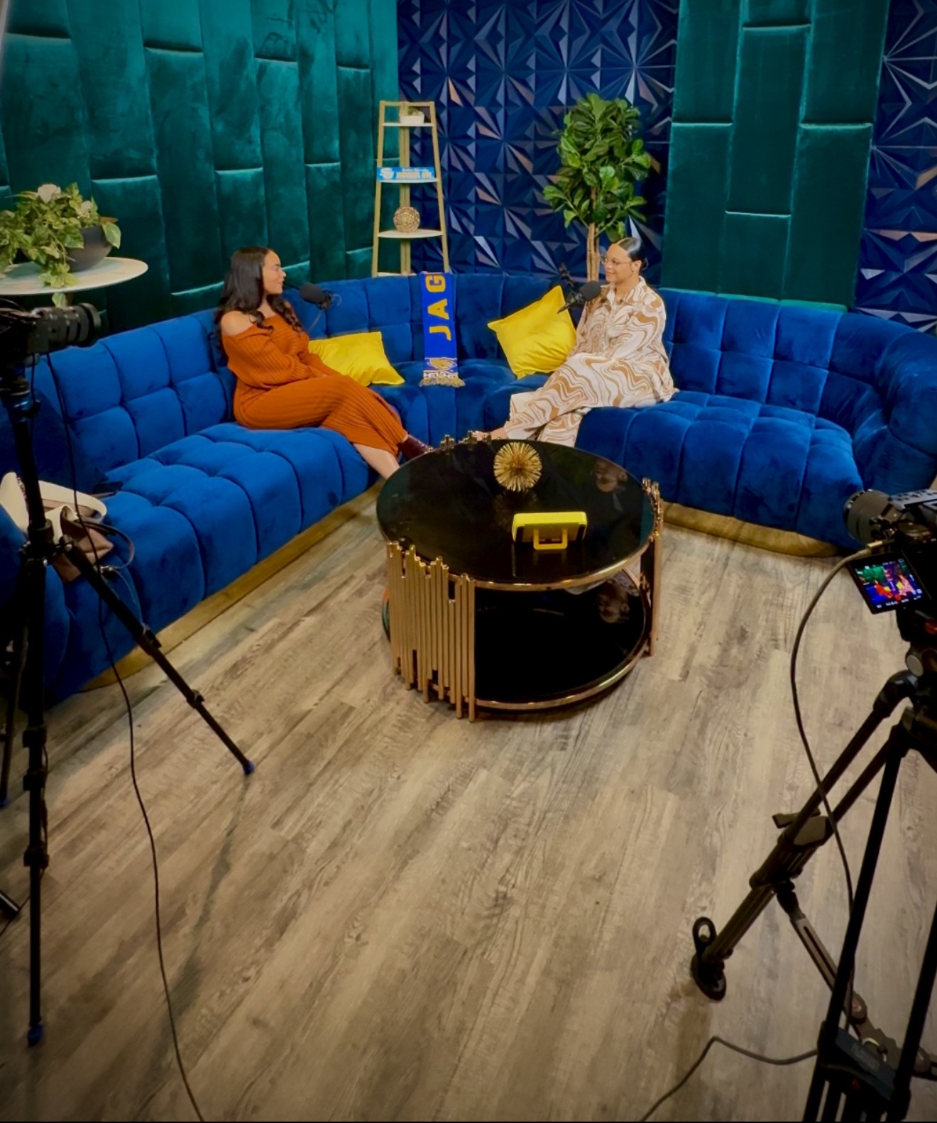 Two women sit on a blue curved sofa in a colorful studio, with cameras and lighting equipment visible.