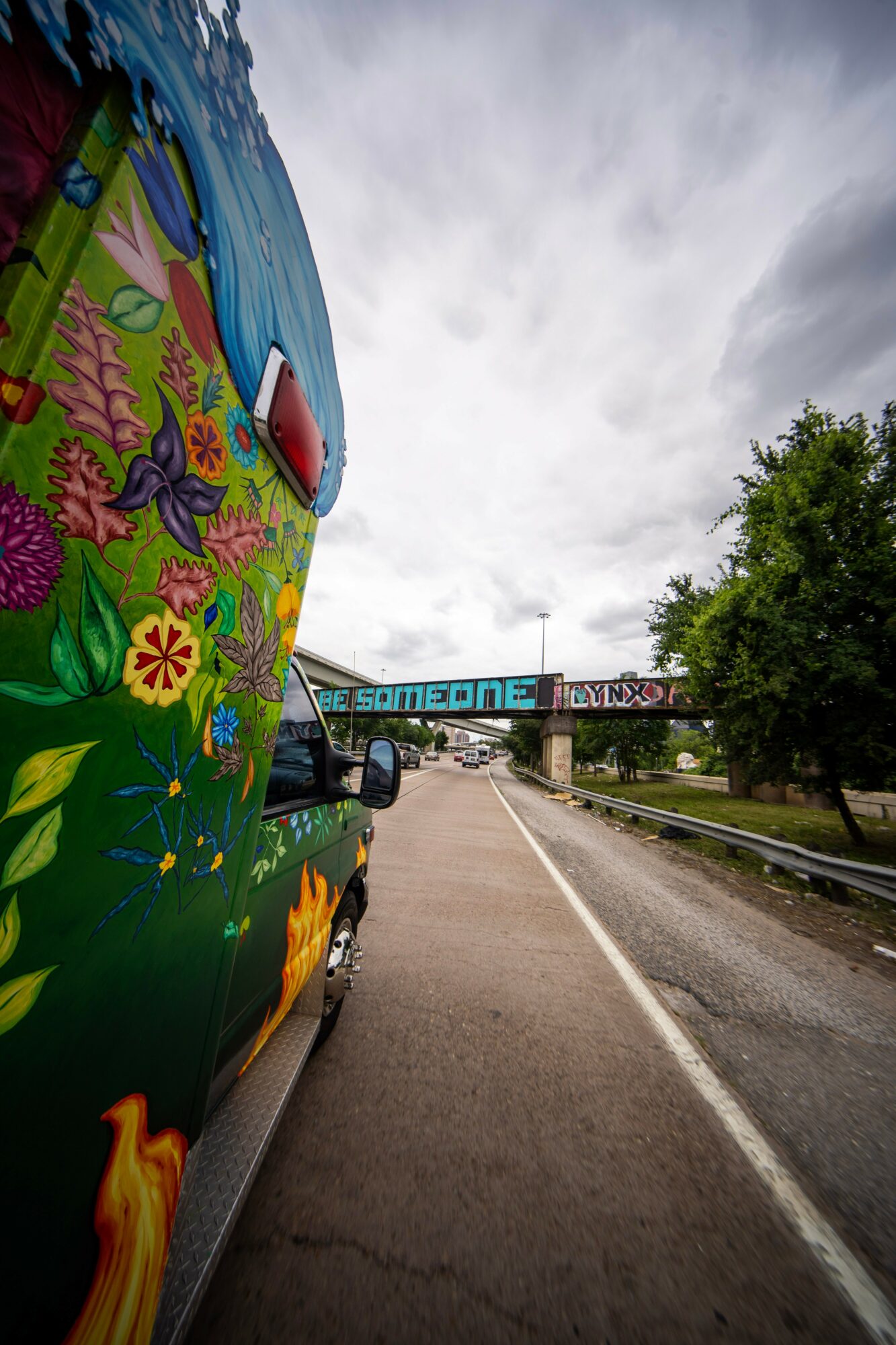 Colorful van with floral and butterfly designs parked on the side of a road, bridge in the background, cloudy sky, trees nearby.