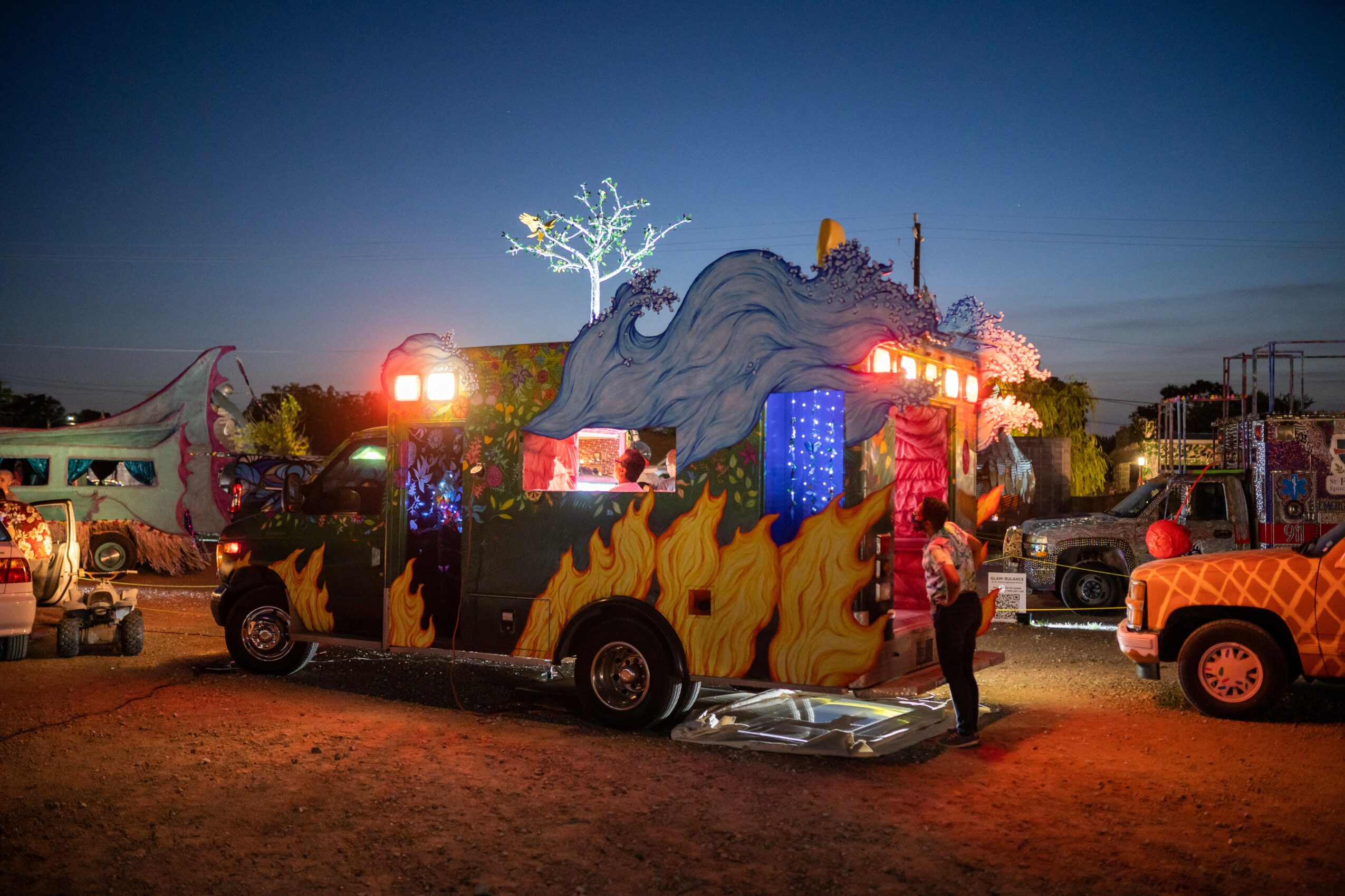 A colorful parade float with a dragon head, flames, and a tree, surrounded by trucks and people at dusk.