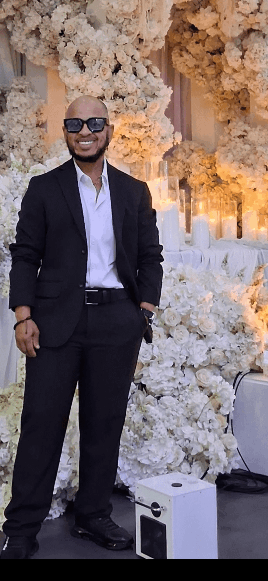 Man in black suit and sunglasses standing in front of floral backdrop at event.