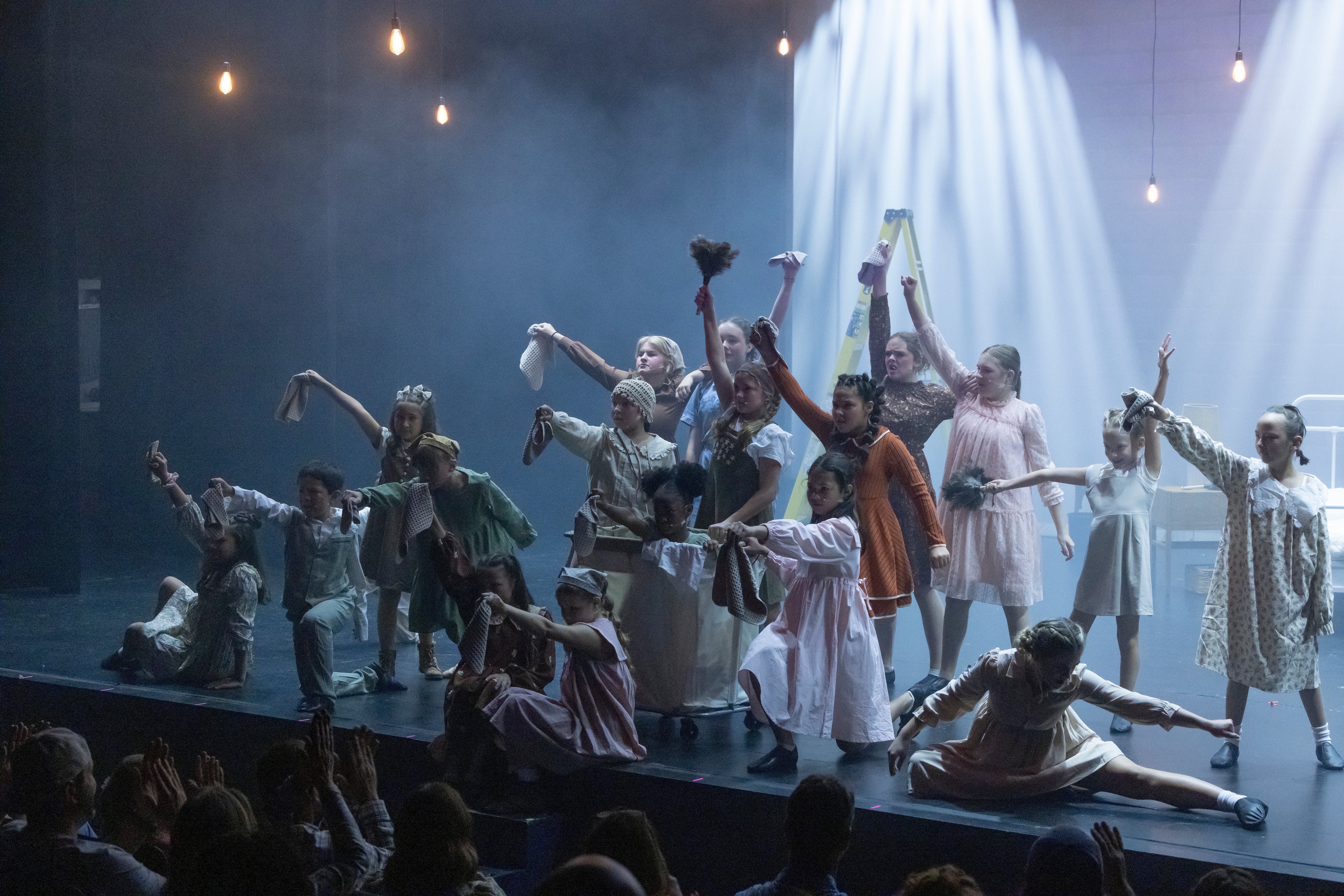 Group of dancers performing on stage with dramatic lighting and flowing costumes, some reaching upward and others in expressive poses.