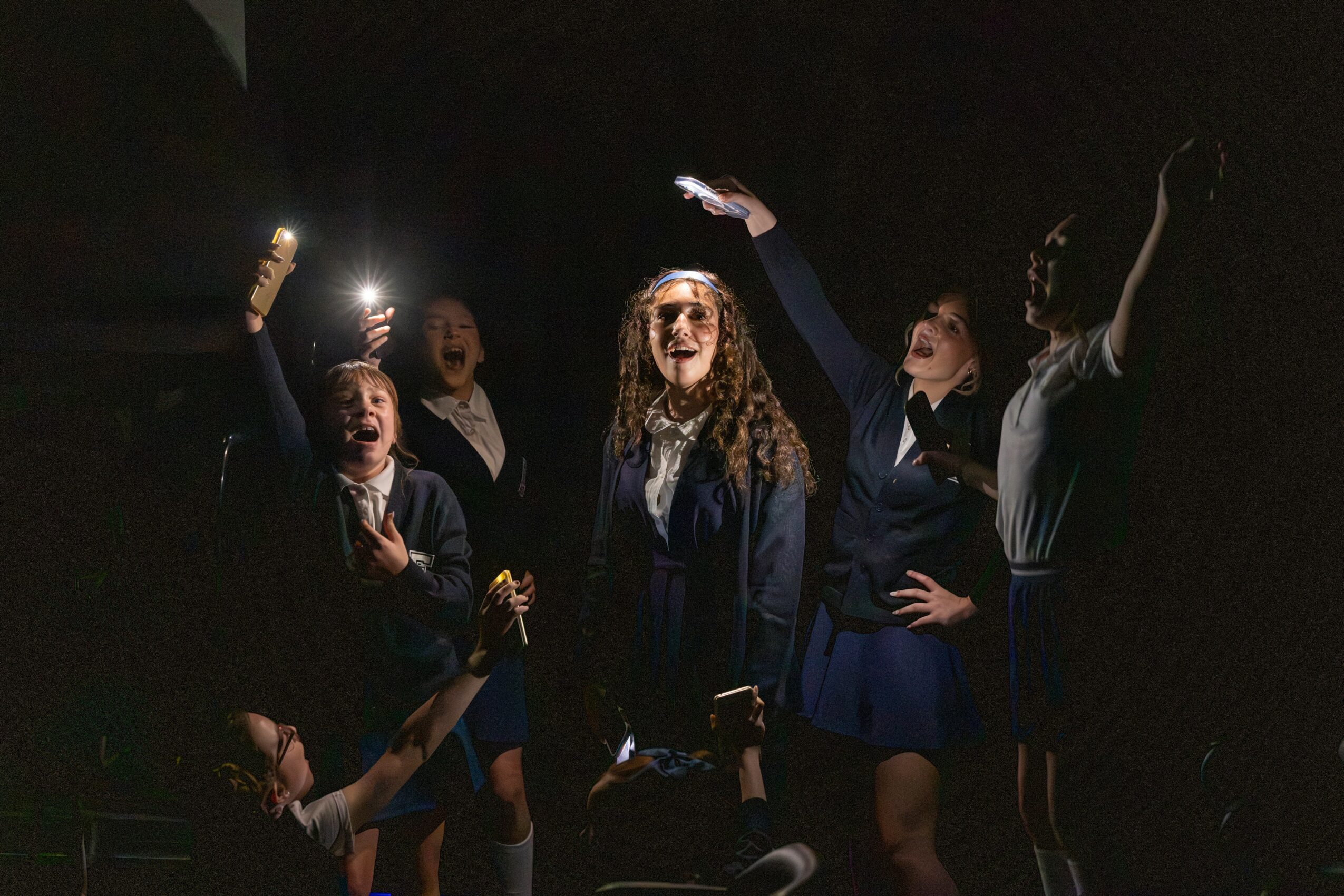Group of five women taking a selfie in a dark setting, some smiling and raising their hands, with one woman in the center.