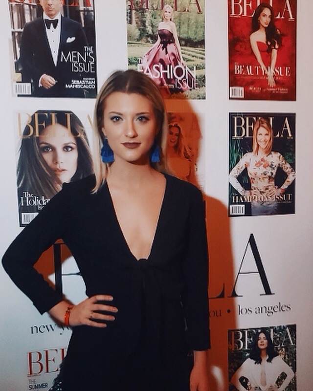 Woman with brown hair in a black dress standing in front of magazine covers on a wall.